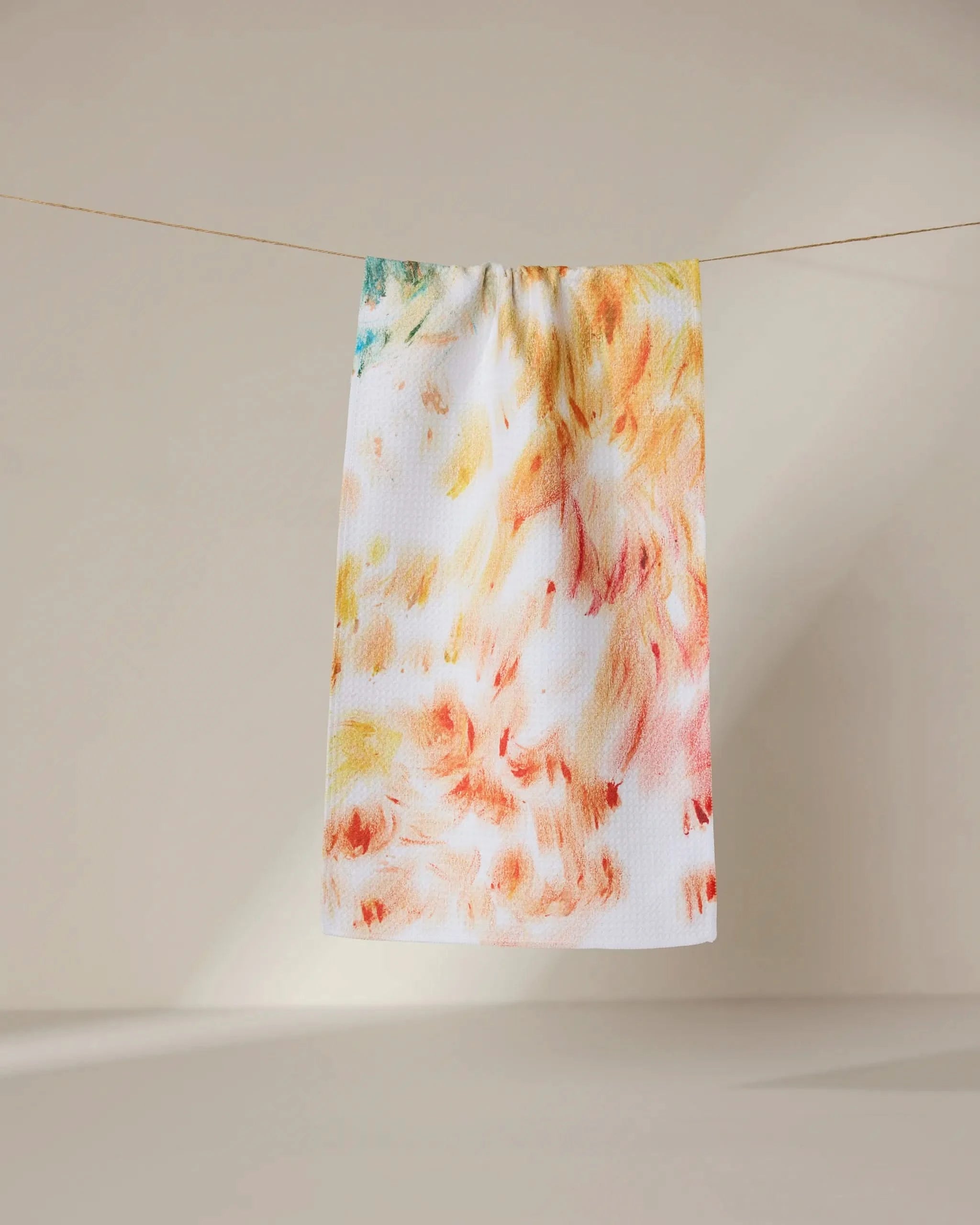 The Waters Edge towel by GEOMETRY, featuring an ultra-absorbent white fabric with an abstract yellow, orange, and red pattern, hangs on a clothesline against a neutral beige background.