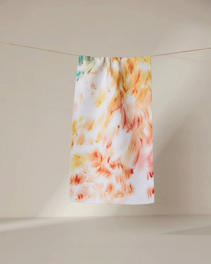 The Waters Edge towel by GEOMETRY, featuring an ultra-absorbent white fabric with an abstract yellow, orange, and red pattern, hangs on a clothesline against a neutral beige background.