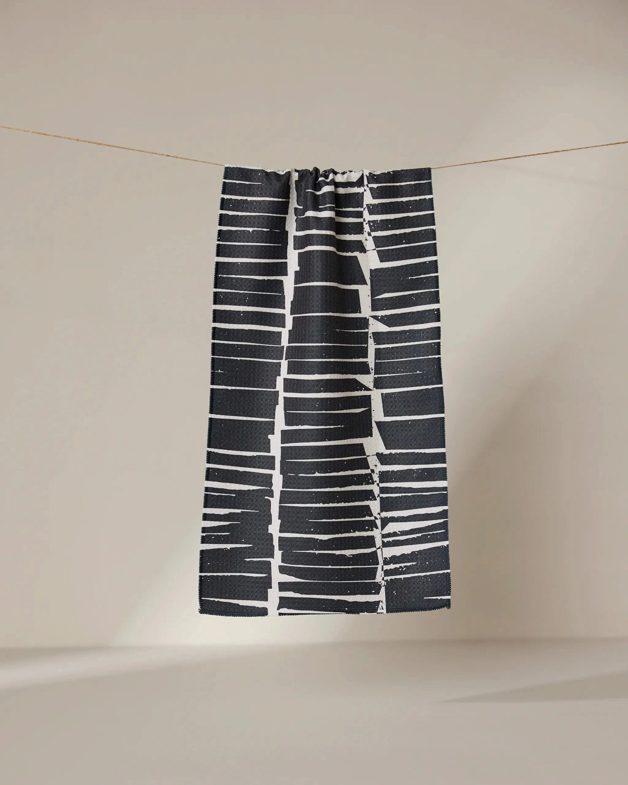 The GEOMETRY Tall Stacks bar towel, crafted from ultra-absorbent recycled materials, features a bold black and white abstract horizontal stripe pattern and hangs on a clothesline against a neutral background.