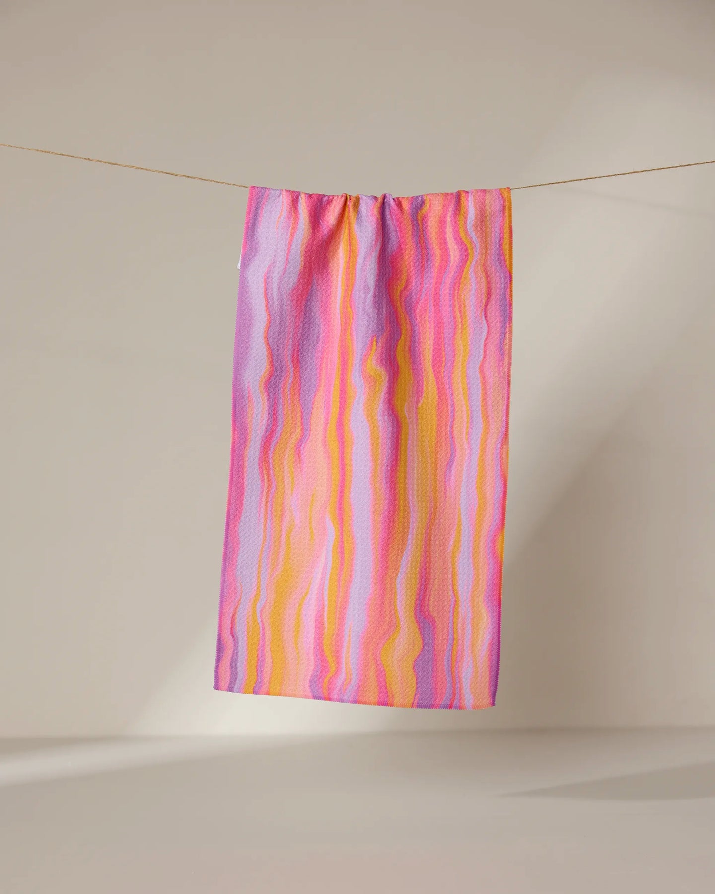 The Rush quick-drying towel by GEOMETRY, featuring wavy pink, yellow, and purple stripes, hangs from a clothesline against a plain beige background.