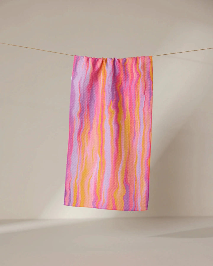The Rush quick-drying towel by GEOMETRY, featuring wavy pink, yellow, and purple stripes, hangs from a clothesline against a plain beige background.