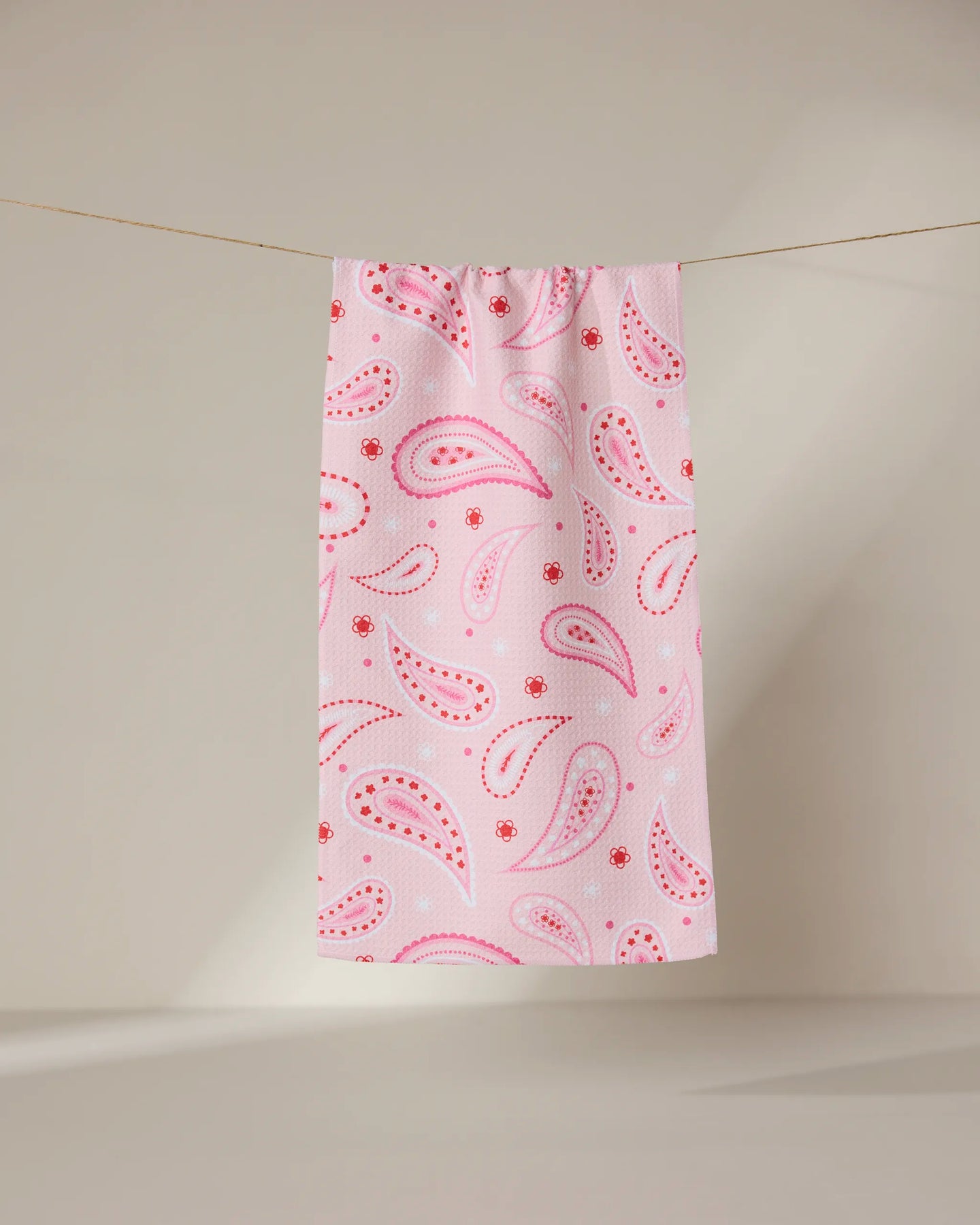 The GEOMETRY Sweet Paisley ultra-absorbent towel in pink, featuring a red and white paisley pattern, hangs on a clothesline against a neutral background.