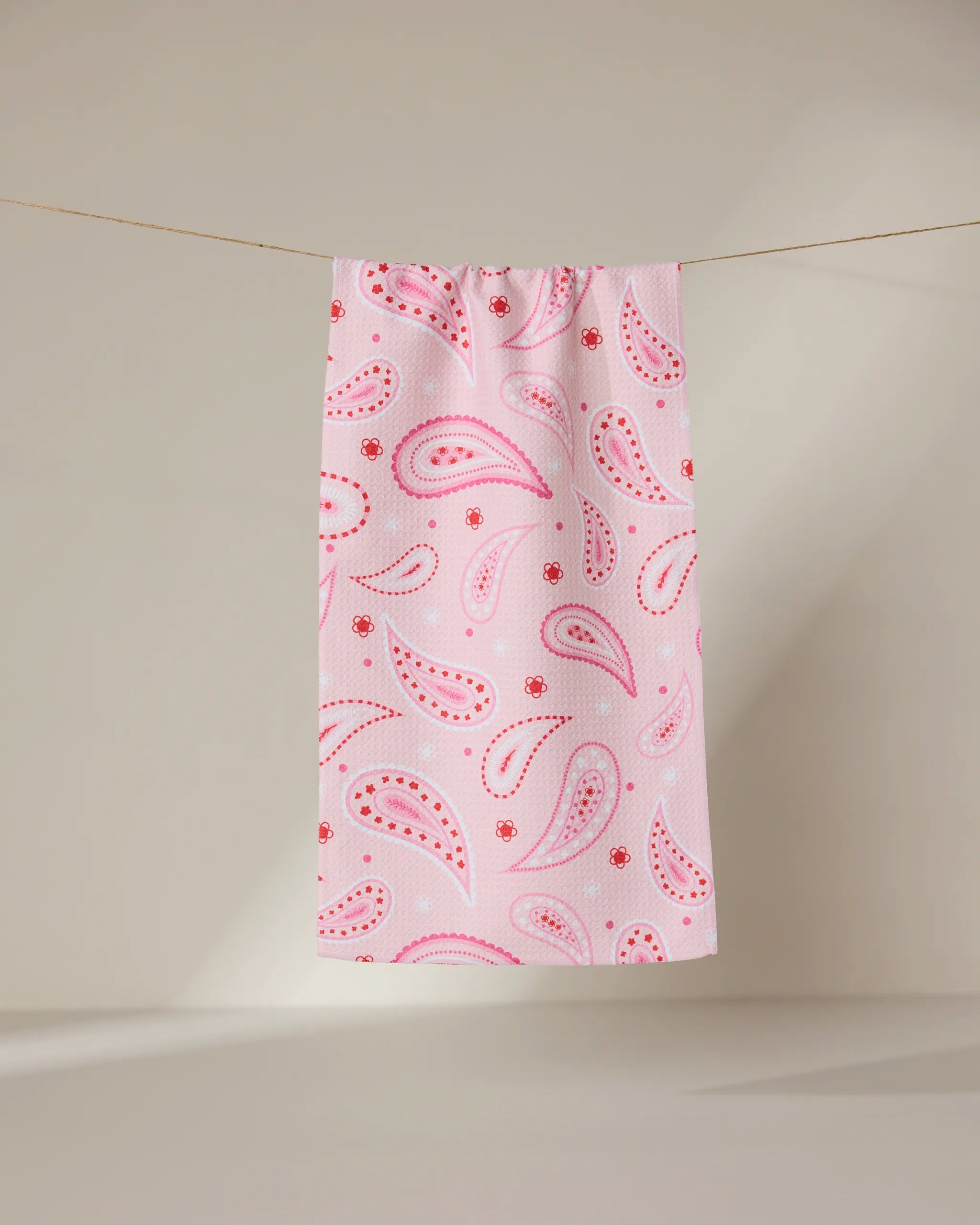 The GEOMETRY Sweet Paisley ultra-absorbent towel in pink, featuring a red and white paisley pattern, hangs on a clothesline against a neutral background.