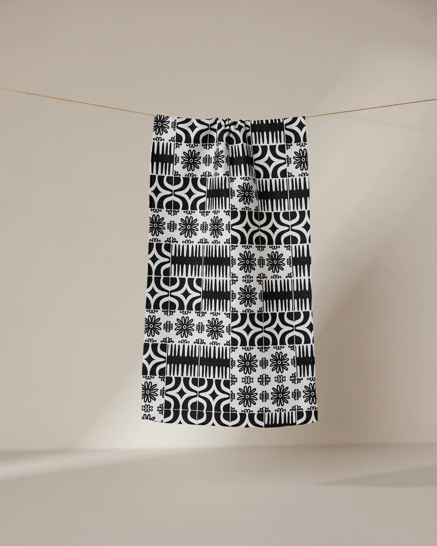 The GEOMETRY Bohemian Mosaic bar towel, crafted from recycled materials, showcases black and white geometric and floral patterns as it hangs from a clothesline against a beige background—a stylish, sustainable choice.
