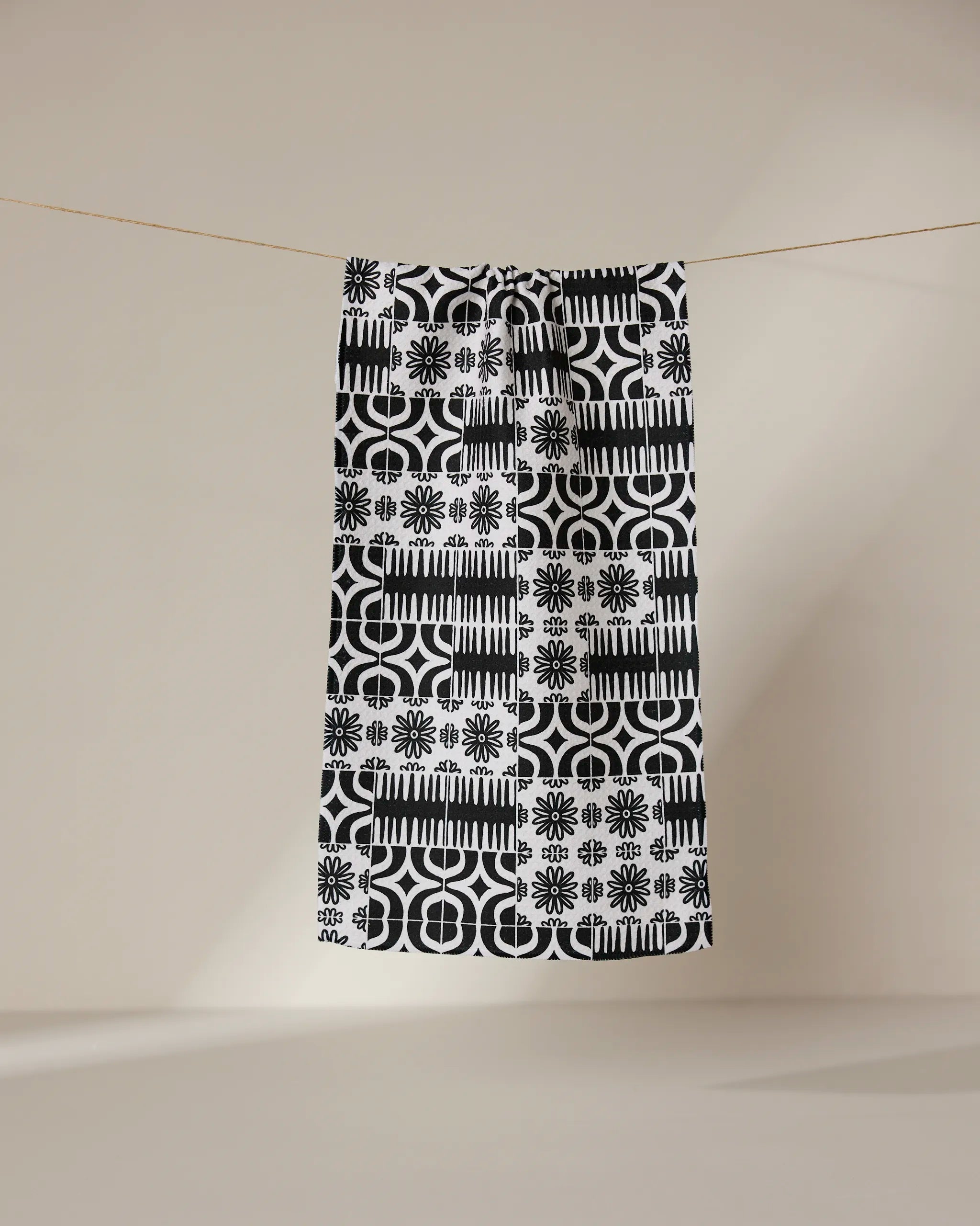 The GEOMETRY Bohemian Mosaic bar towel, crafted from recycled materials, showcases black and white geometric and floral patterns as it hangs from a clothesline against a beige background—a stylish, sustainable choice.