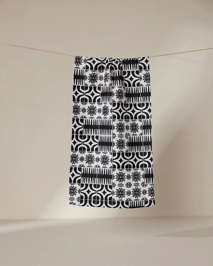 The GEOMETRY Bohemian Mosaic bar towel, crafted from recycled materials, showcases black and white geometric and floral patterns as it hangs from a clothesline against a beige background—a stylish, sustainable choice.