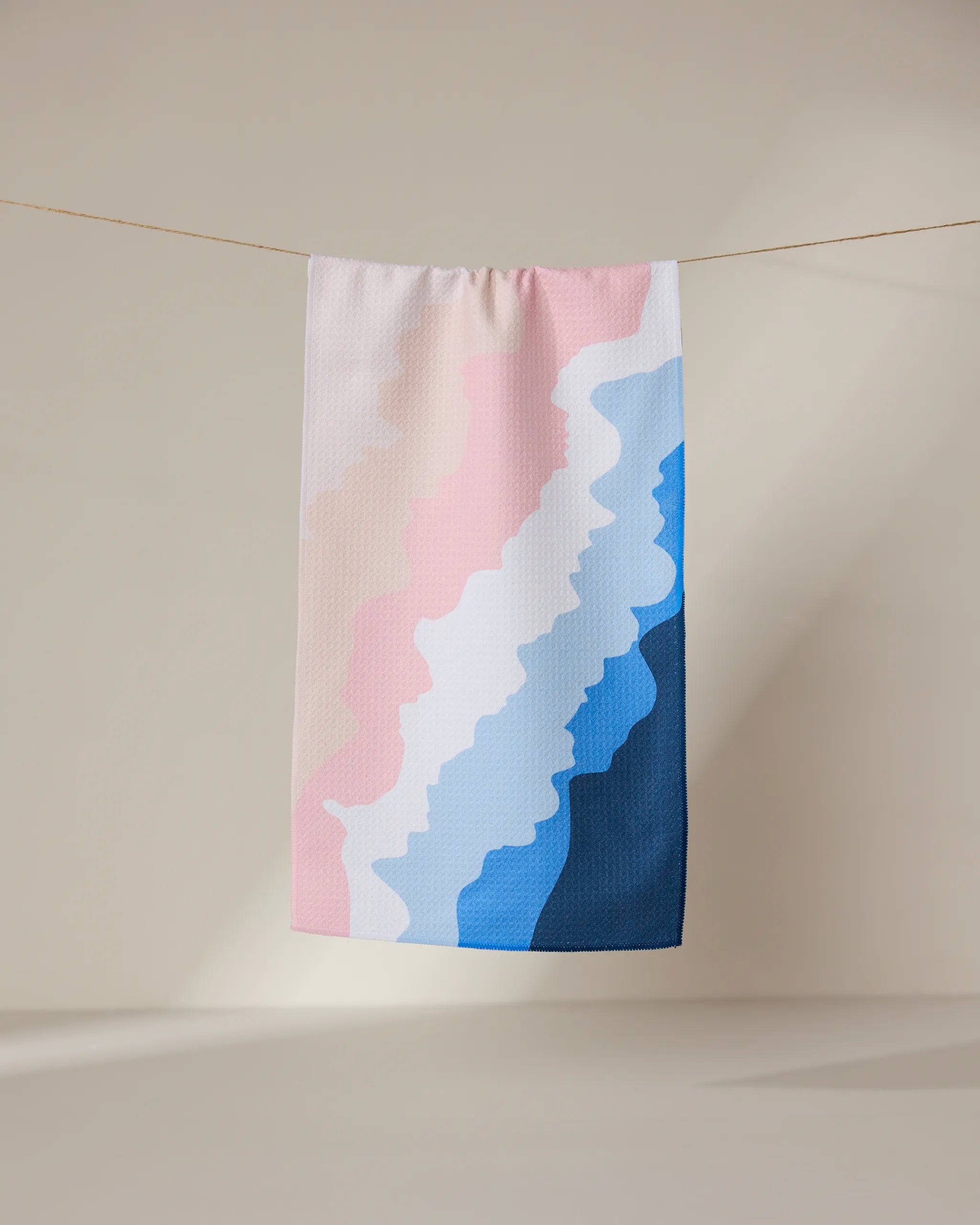 The GEOMETRY Shore towel, featuring a wavy abstract pattern in pink, white, and blue, hangs from a line against a neutral beige background. Ultra-absorbent and stylish, it combines function with modern design.