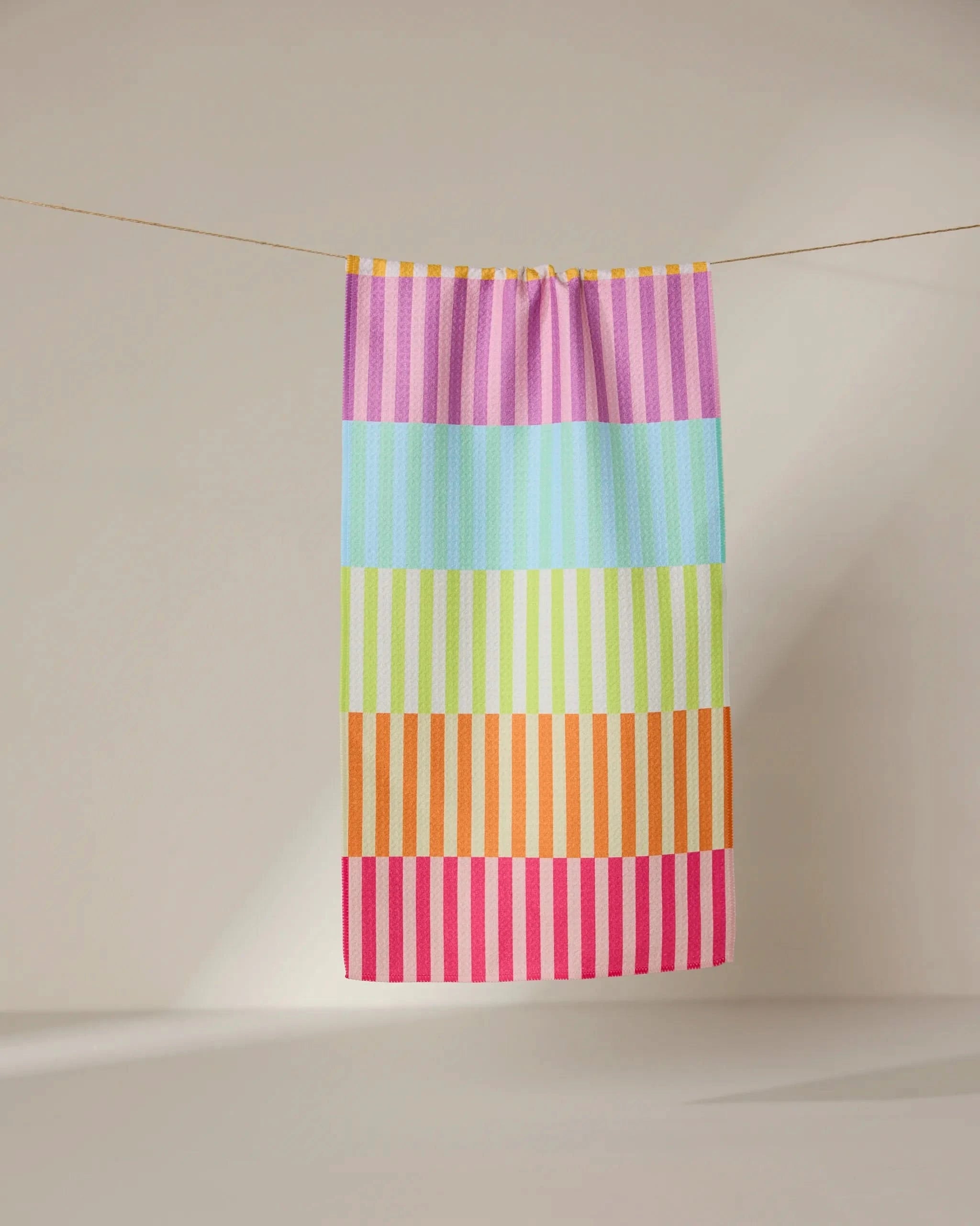 The GEOMETRY Neon Nights towel, featuring ultra-absorbent fabric and horizontal pastel stripes in pink, purple, blue, green, yellow, orange, and red, hangs on a clothesline against a neutral background.
