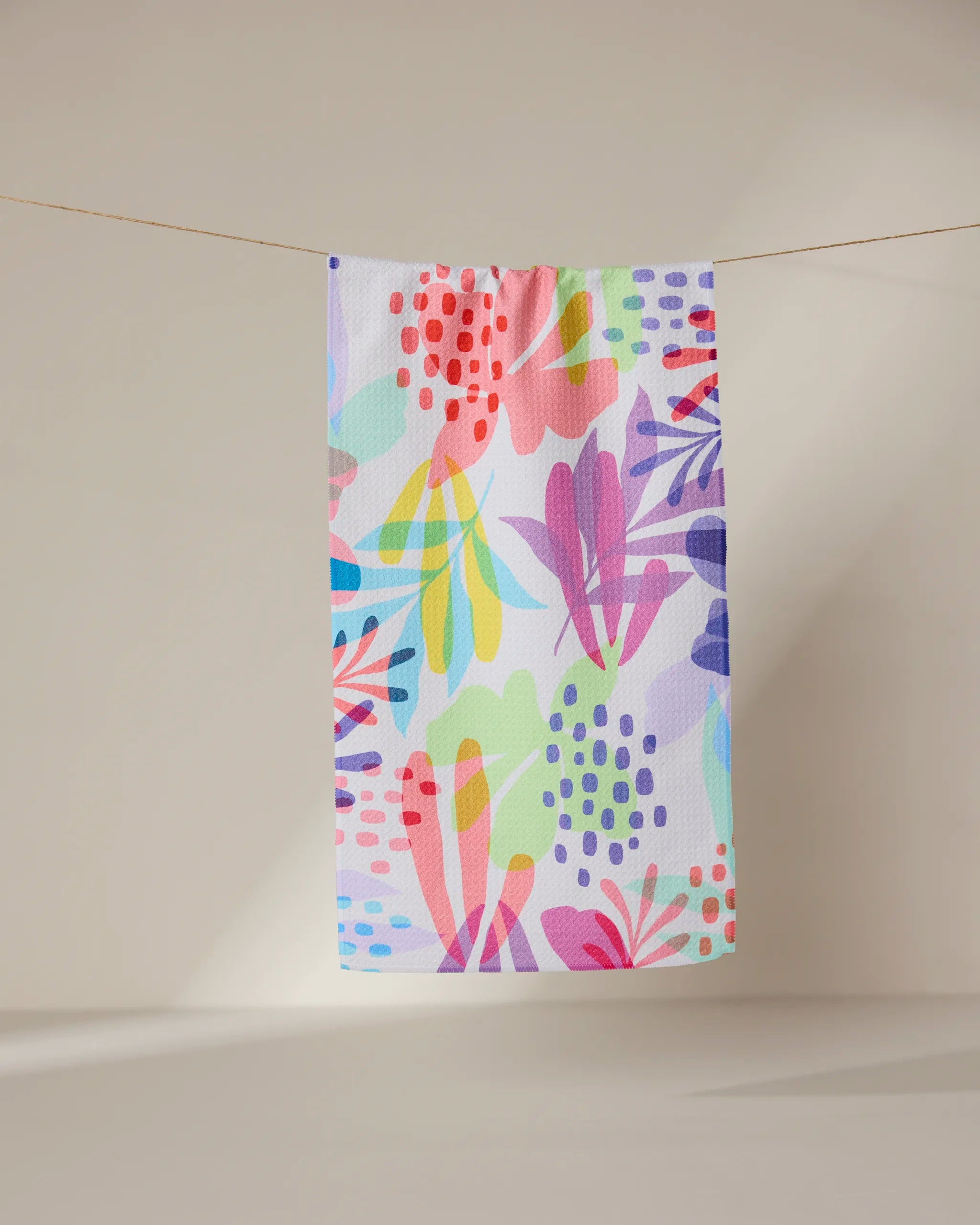 The GEOMETRY Botanical Burst towel, featuring a vibrant abstract floral pattern in pink, purple, yellow, blue, and green, hangs on a clothesline against a neutral, softly lit background.