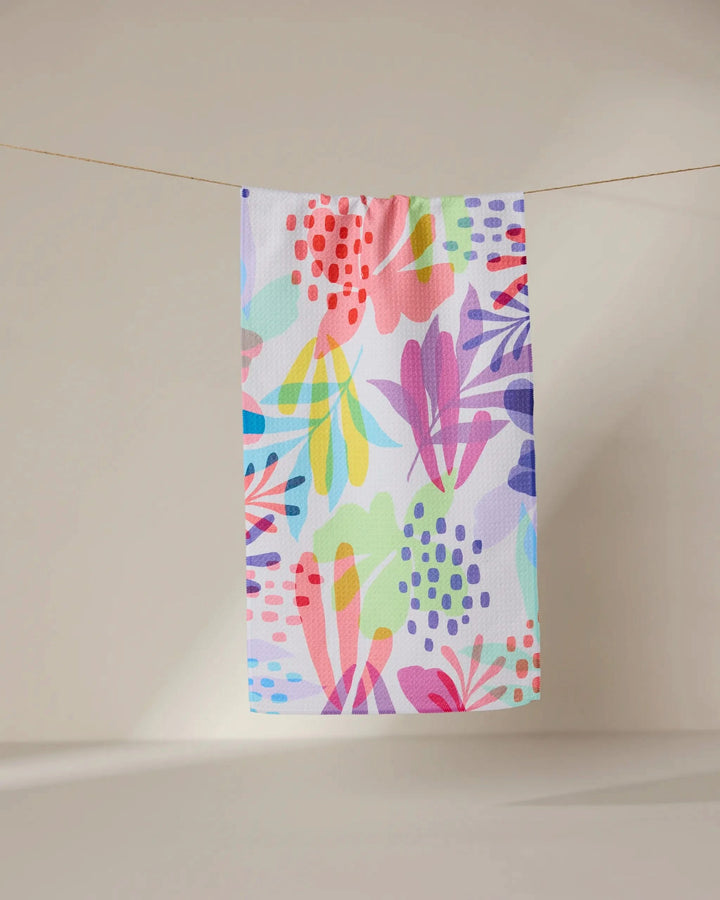 The GEOMETRY Botanical Burst towel, featuring a vibrant abstract floral pattern in pink, purple, yellow, blue, and green, hangs on a clothesline against a neutral, softly lit background.
