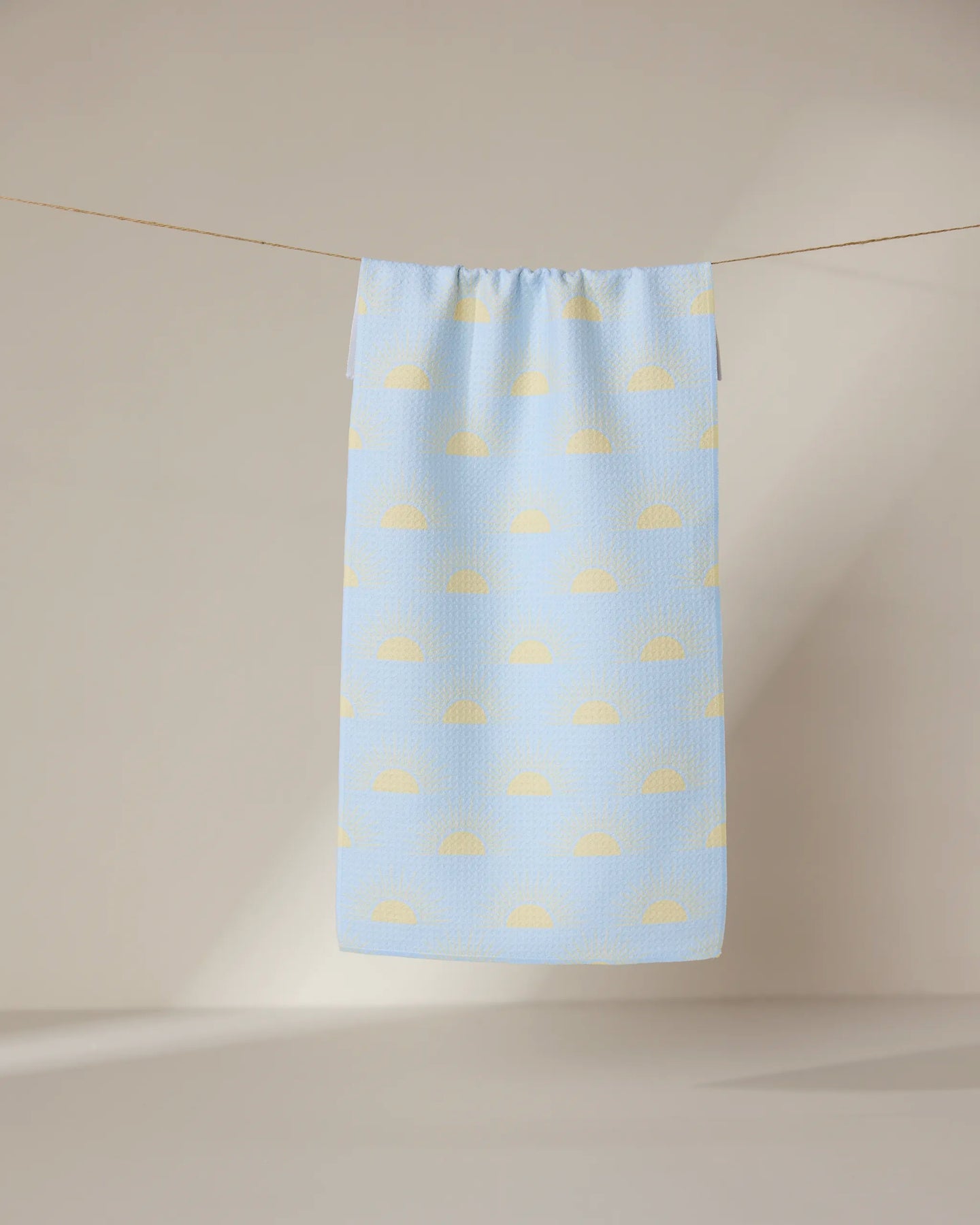 The GEOMETRY Soft Summer Sun towel, ultra-absorbent and light blue with a pattern of yellow semicircles, hangs on a clothesline against a softly lit neutral background.