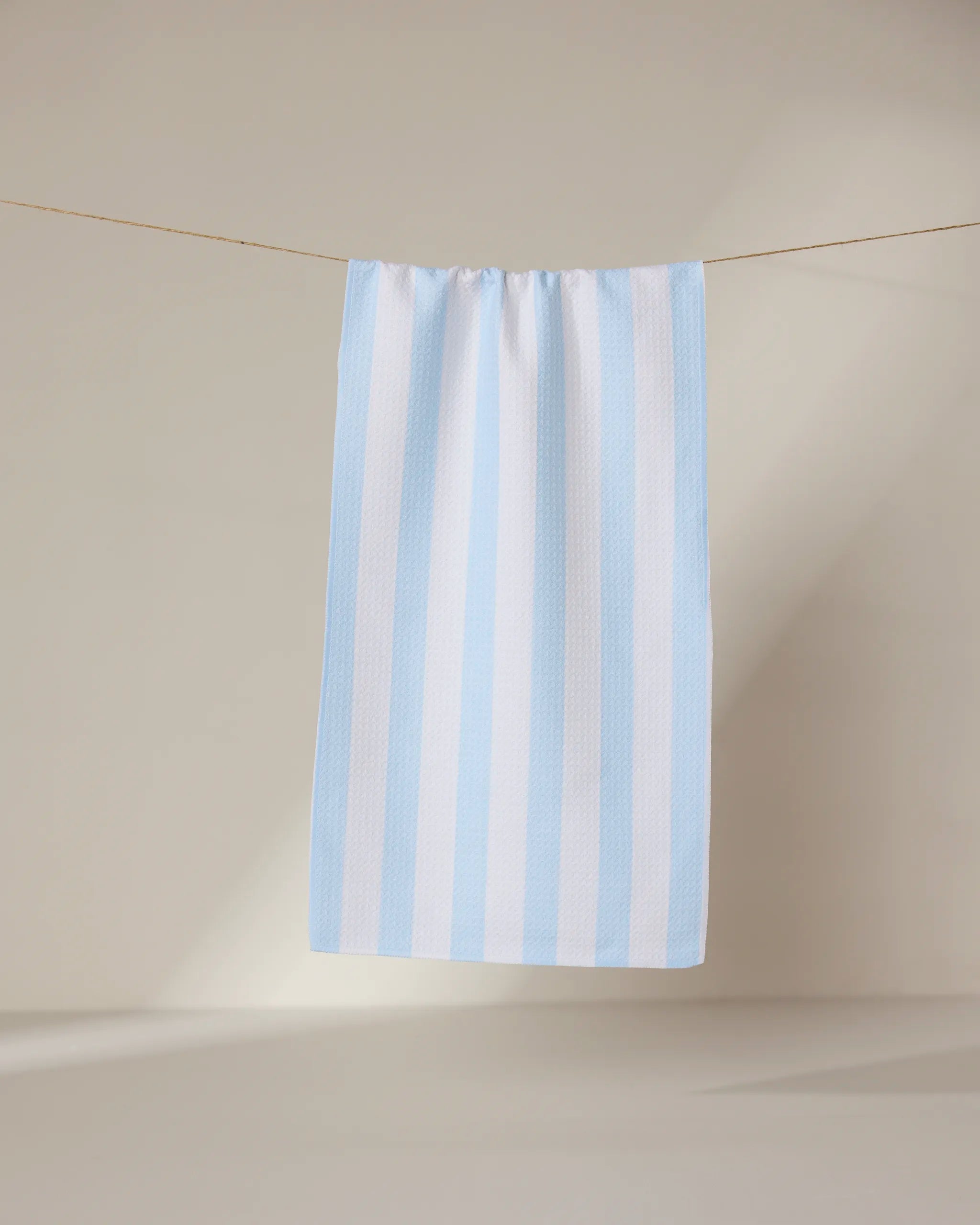 The GEOMETRY Seaside Stripes recycled kitchen towel, featuring light blue and white vertical stripes, hangs neatly on a clothesline against a neutral wall with soft lighting.