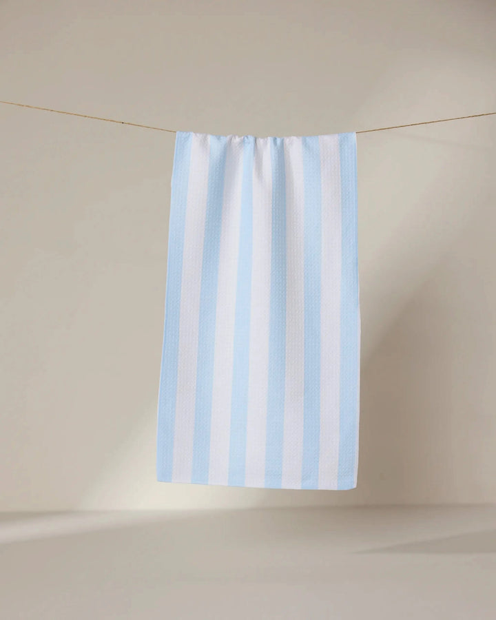 The GEOMETRY Seaside Stripes recycled kitchen towel, featuring light blue and white vertical stripes, hangs neatly on a clothesline against a neutral wall with soft lighting.