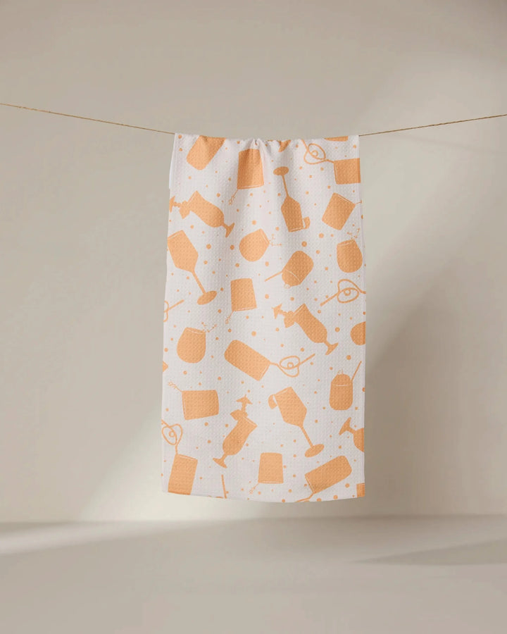 The GEOMETRY Bach Brunch bar towel is white, made from recycled materials, and features an orange hairdressing tool pattern. It hangs on a clothesline against a neutral background.