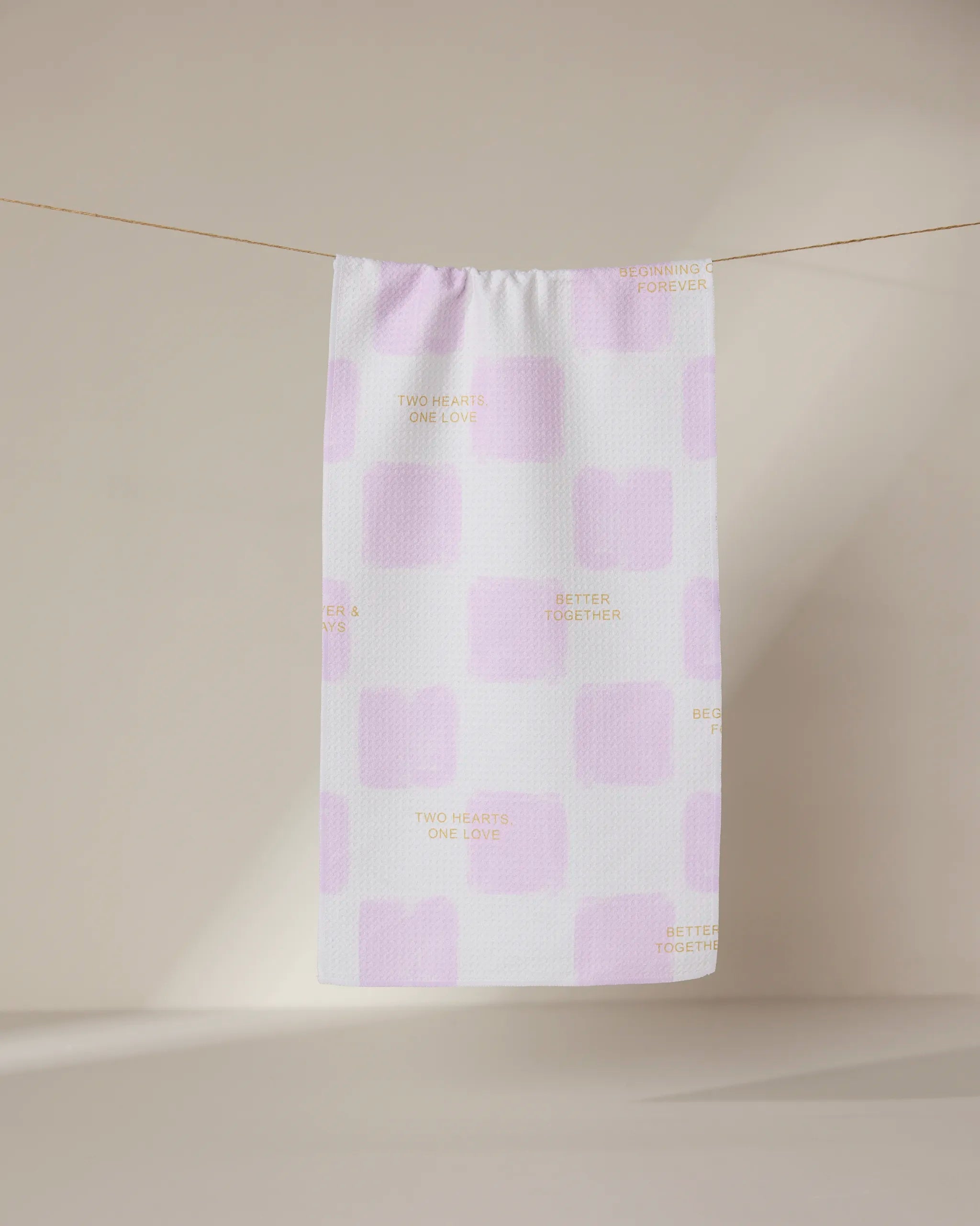 The GEOMETRY You and I towel, featuring a white base with pale pink squares and light brown phrases like TWO HEARTS ONE LOVE and BETTER TOGETHER, hangs on a clothesline against a neutral background.