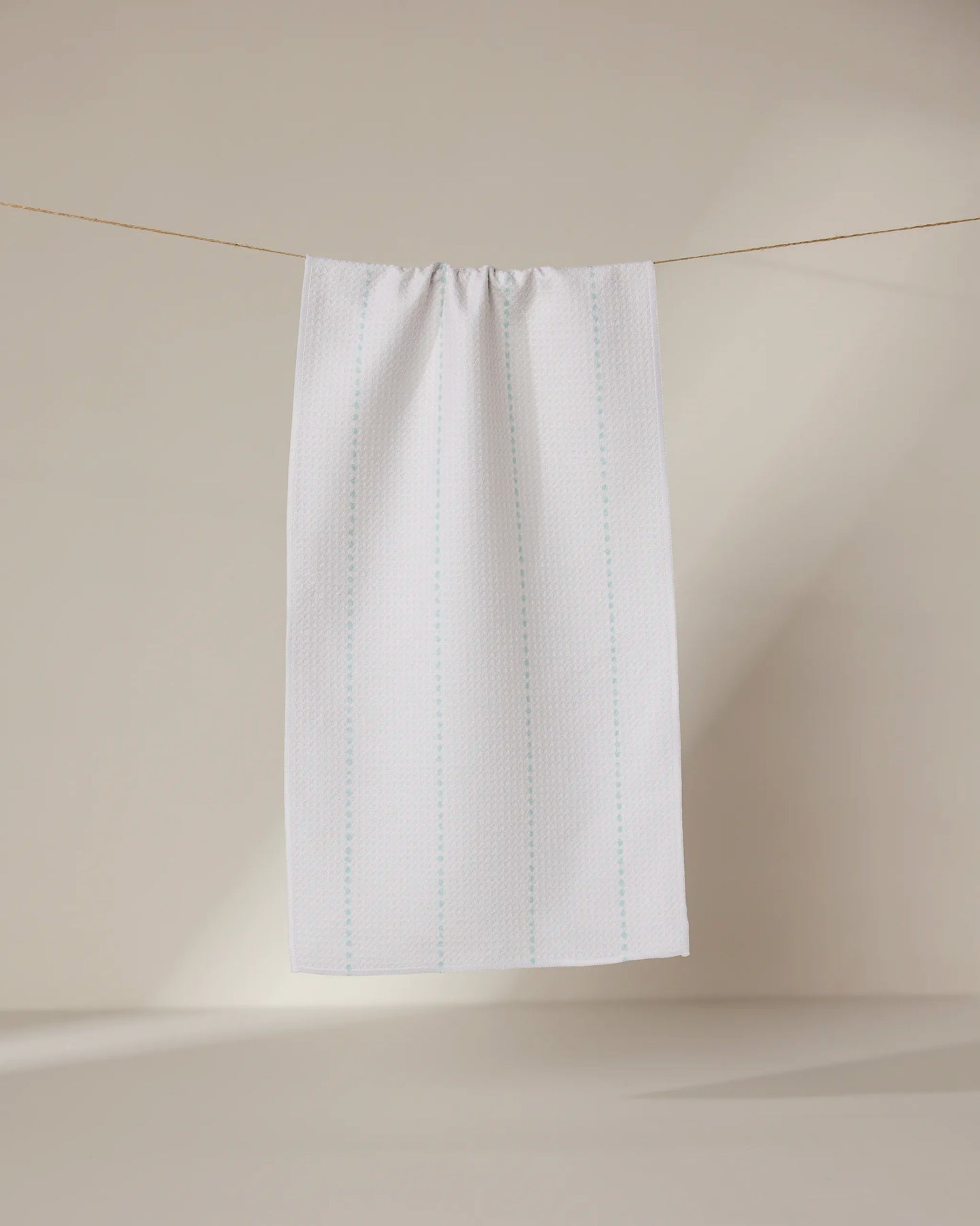 The GEOMETRY Dotted Dunes towel, featuring ultra-absorbent fabric with subtle blue vertical lines on white, hangs on a thin string against a beige backdrop—an eco-friendly and stylish choice for any bar.