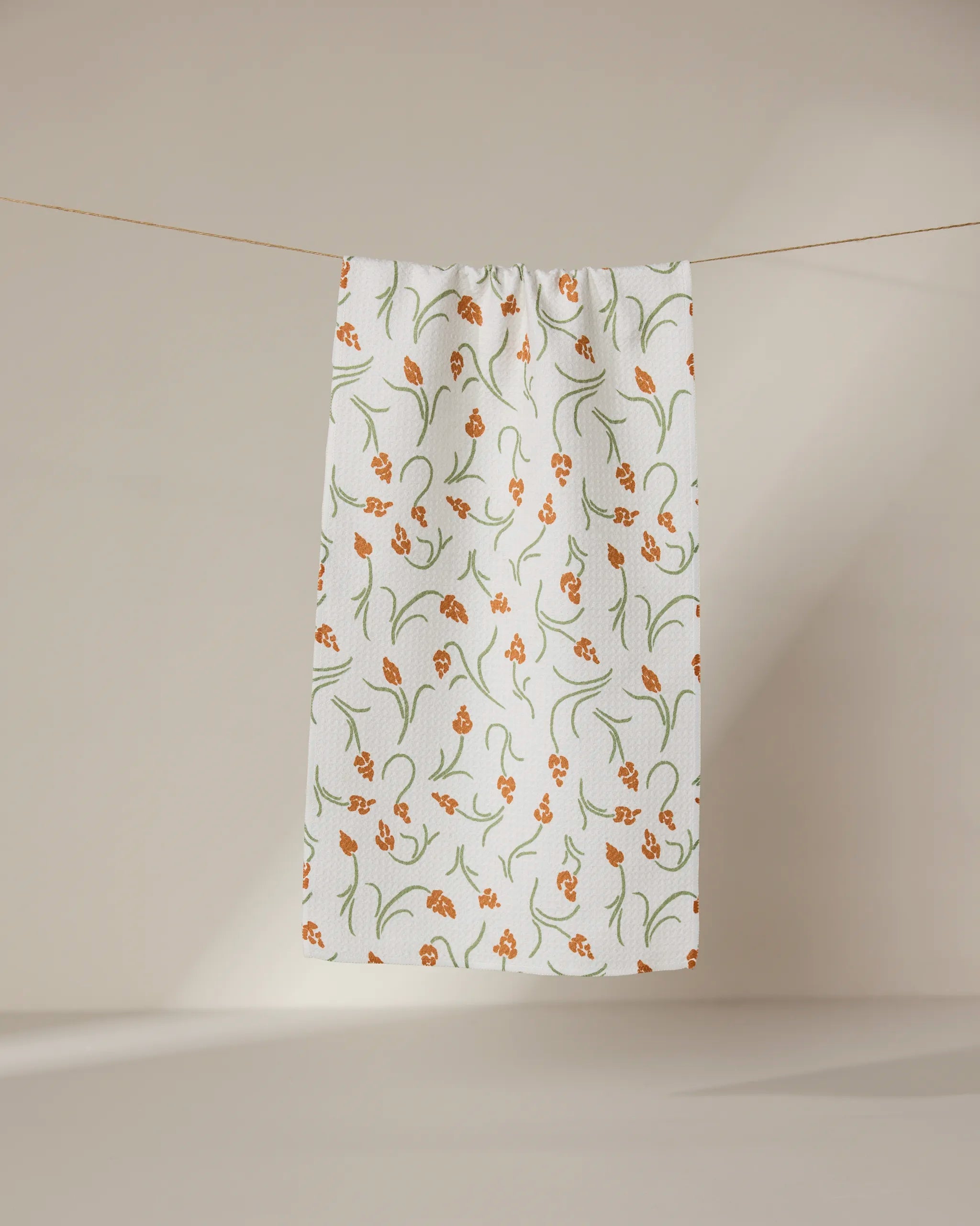 The GEOMETRY Crayon Floral ultra-absorbent white towel, featuring an orange and green floral design, hangs against a neutral beige background.