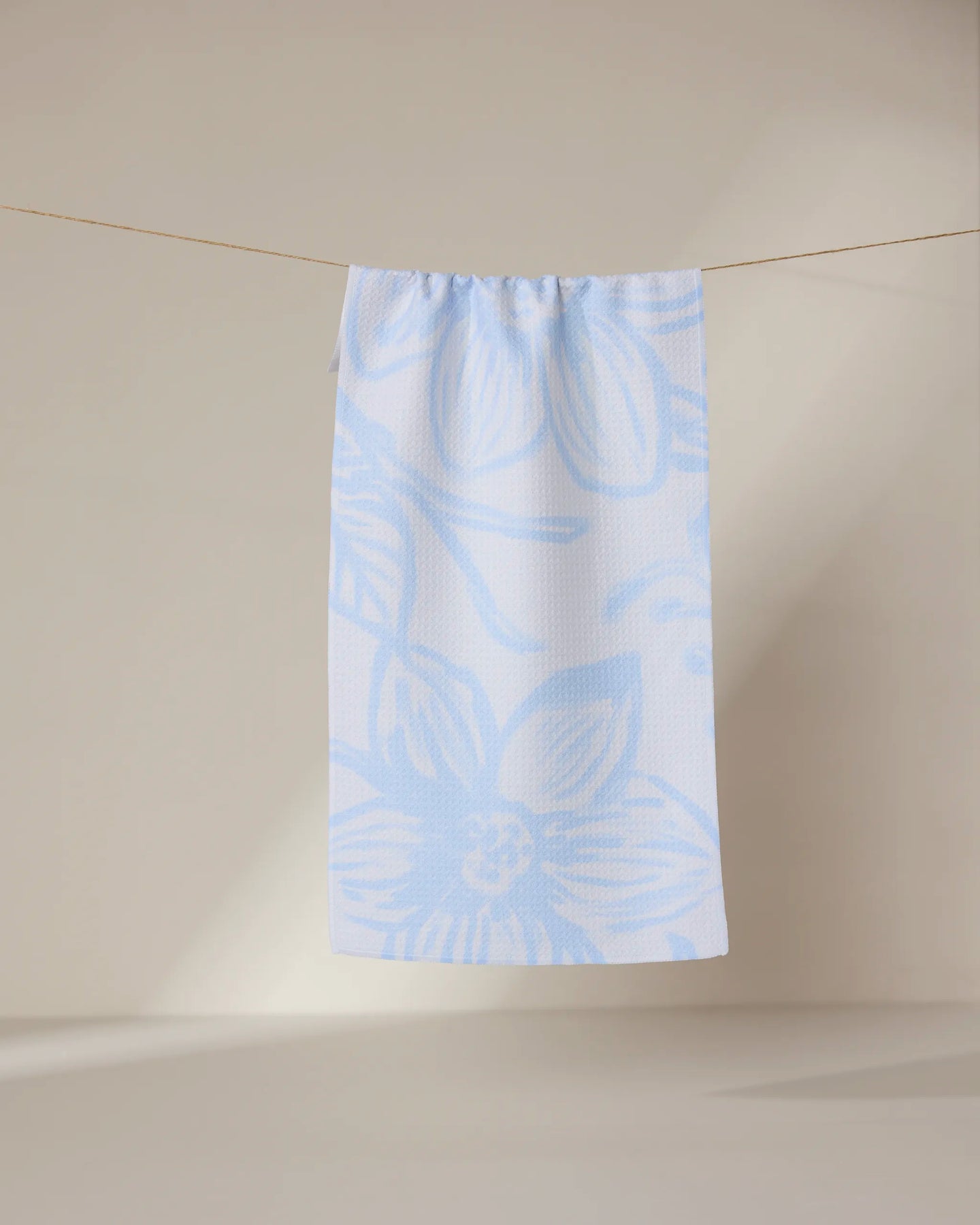 The GEOMETRY Something Blue bar towel, featuring light blue floral patterns, hangs on a thin clothesline against a beige background with soft light casting gentle shadows on the wall and floor.