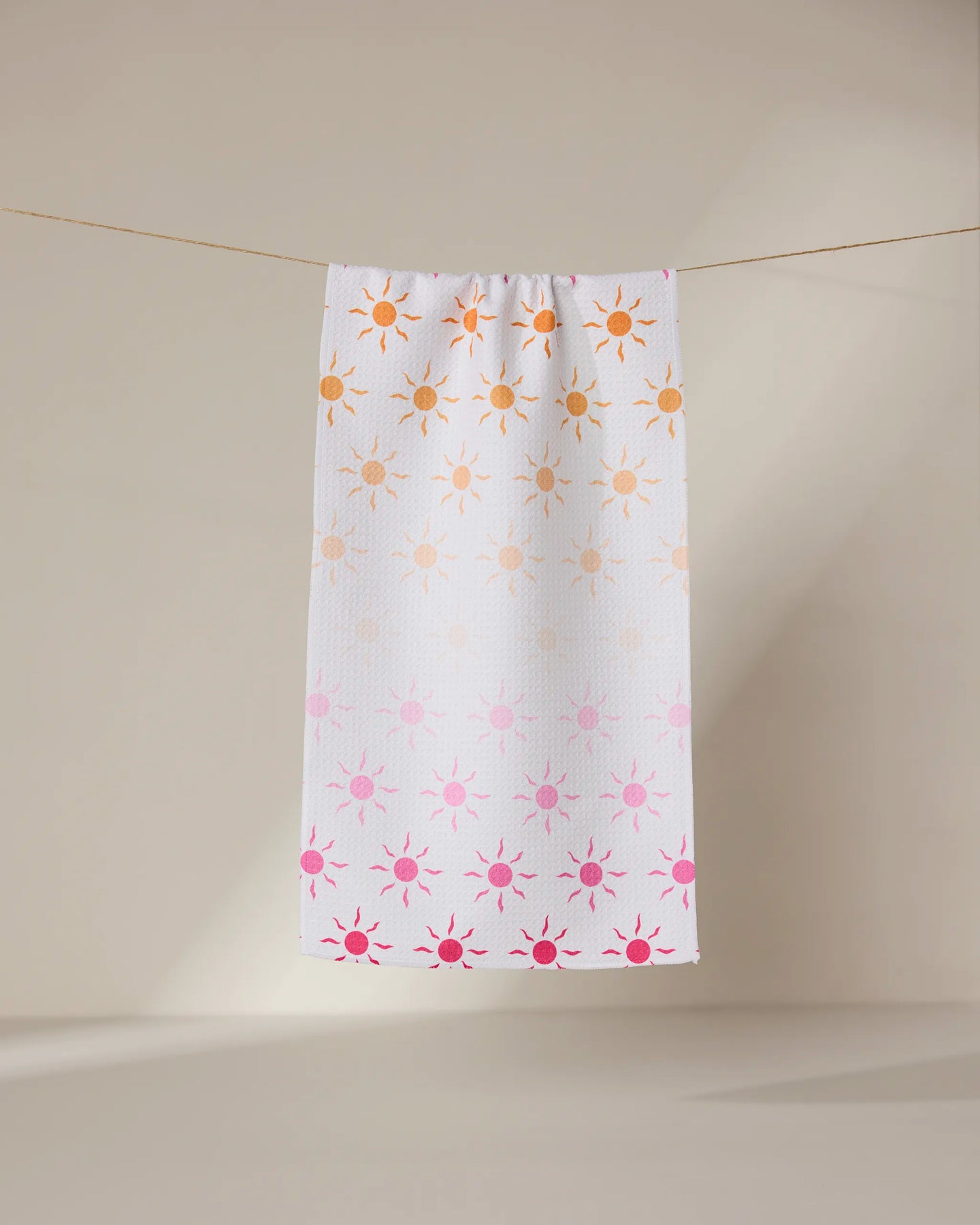 The GEOMETRY Fun in the Sun bar towel, featuring orange and pink sun patterns on white, hangs on a thin clothesline against a beige background.