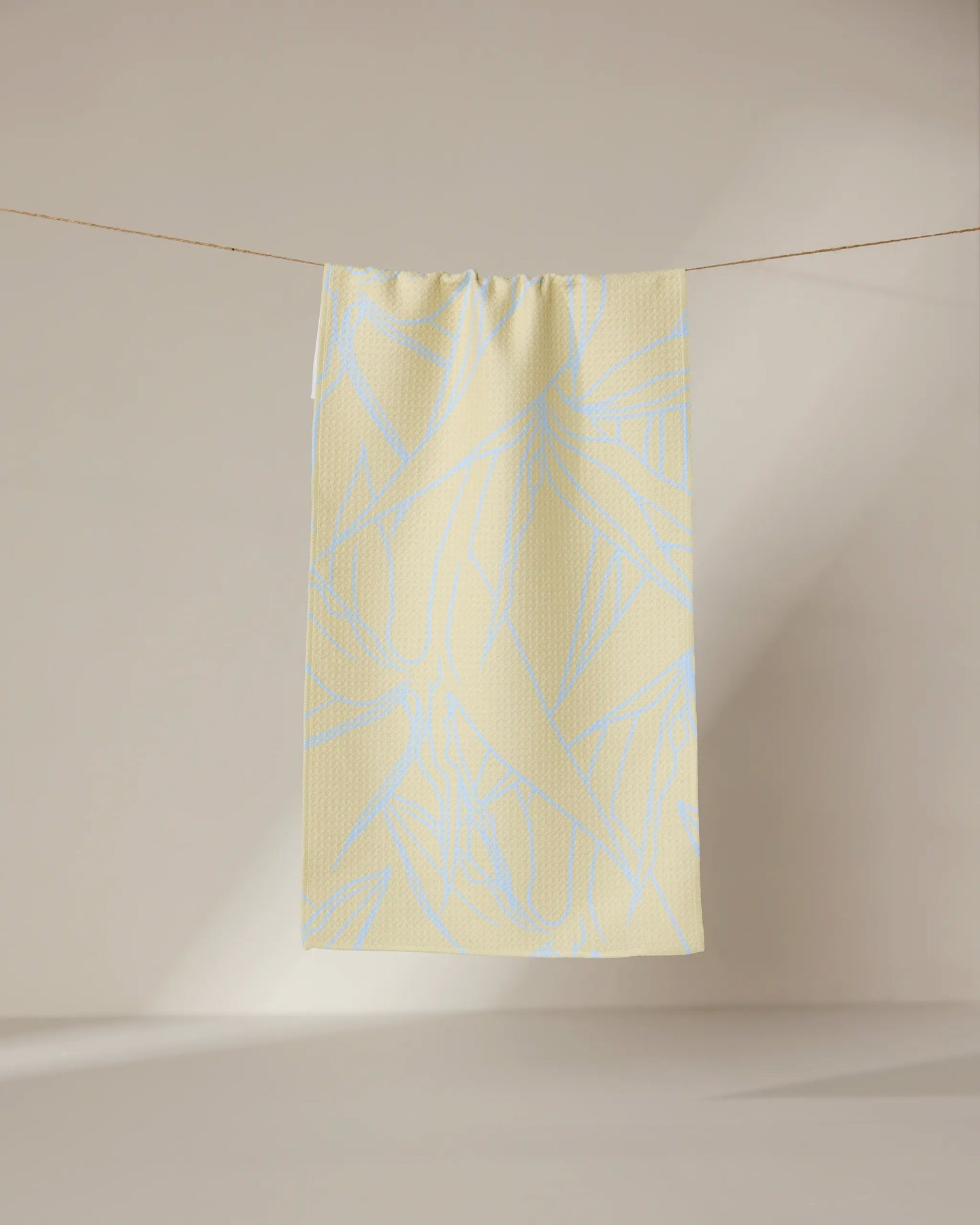 The GEOMETRY Paradise Blooms towel, featuring a pale blue abstract leaf design on light yellow, hangs neatly on a thin clothesline against a softly lit, neutral background.