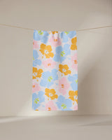 The GEOMETRY Hula Hues bar towel, featuring vibrant blue, yellow, green, and pink florals, hangs on a clothesline against a neutral beige background. Made from ultra-absorbent recycled materials.