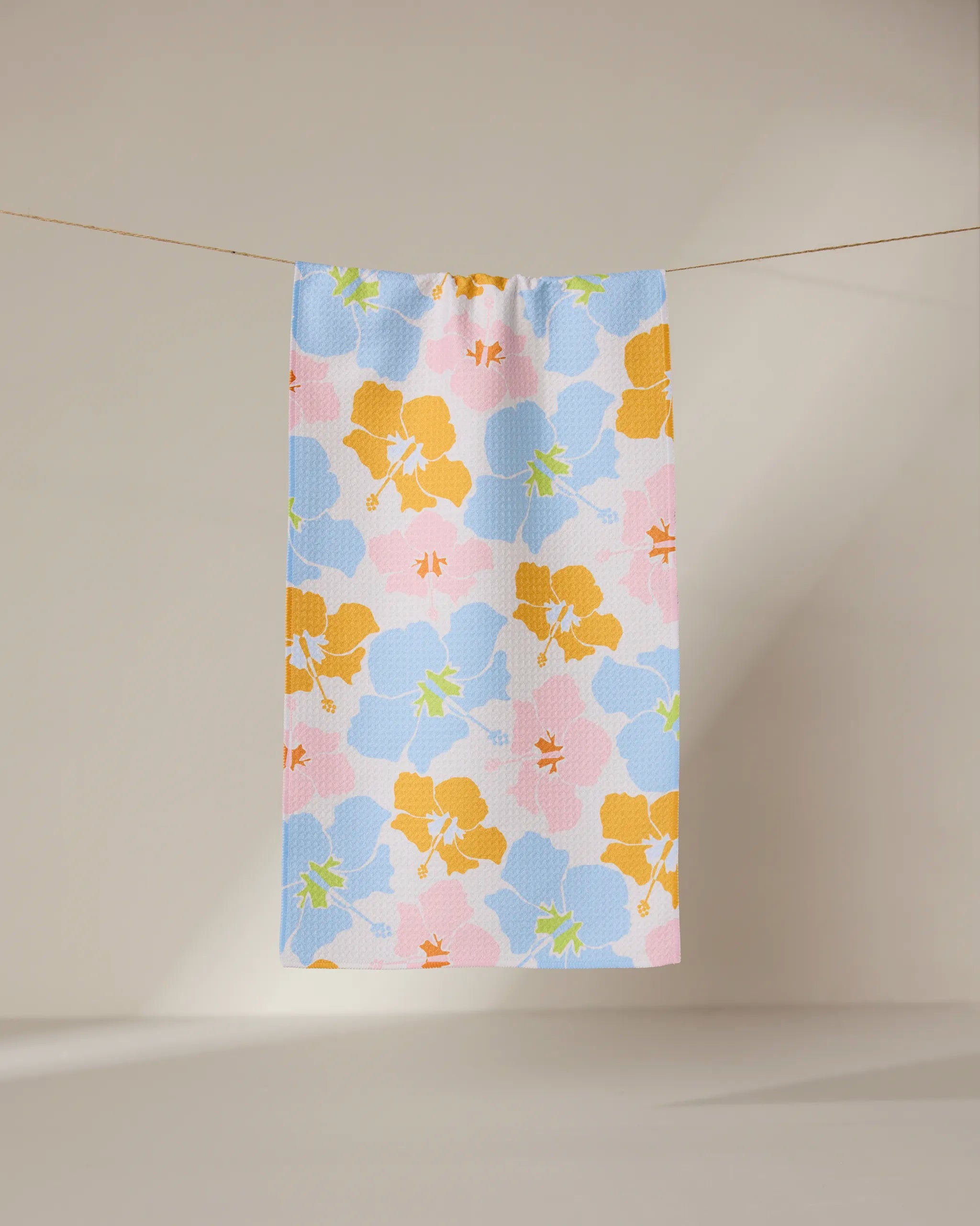 The GEOMETRY Hula Hues bar towel, featuring vibrant blue, yellow, green, and pink florals, hangs on a clothesline against a neutral beige background. Made from ultra-absorbent recycled materials.