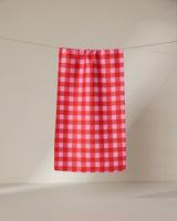 The GEOMETRY Cherry Gingham ultra-absorbent towel in red and pink checks hangs neatly on a clothesline against a plain, light background.