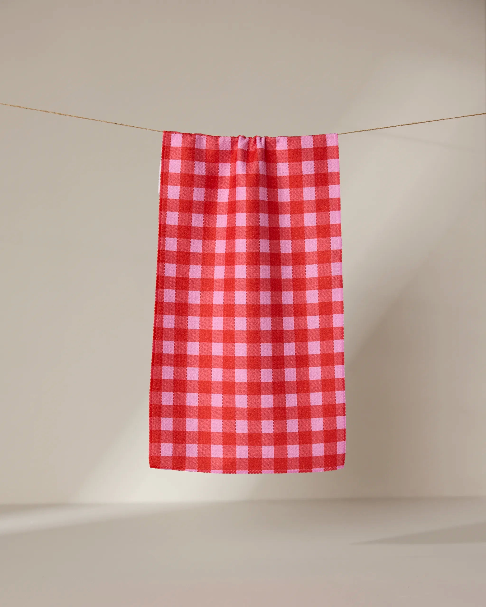 The GEOMETRY Cherry Gingham ultra-absorbent towel in red and pink checks hangs neatly on a clothesline against a plain, light background.