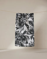 The GEOMETRY Caring towel, white and quick-drying with a bold black floral pattern of oversized flowers and leaves, hangs on a clothesline against a neutral beige background.