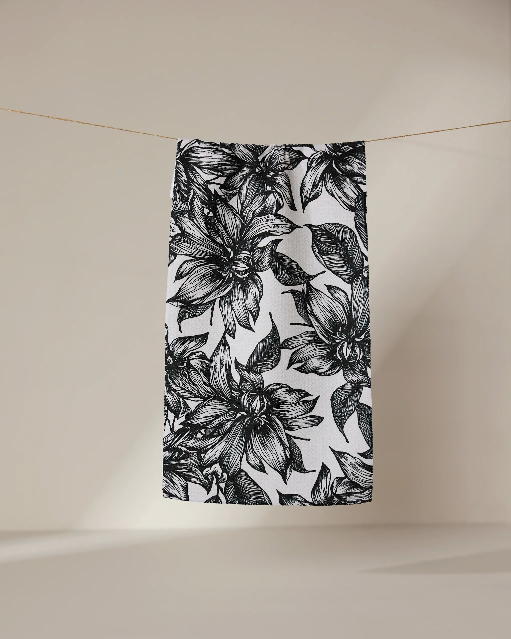 The GEOMETRY Caring towel, white and quick-drying with a bold black floral pattern of oversized flowers and leaves, hangs on a clothesline against a neutral beige background.