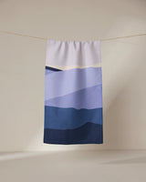 The GEOMETRY Mountainous rectangular towel, ultra-absorbent and adorned with blue and purple mountain-inspired gradients, hangs from a string against a neutral background.