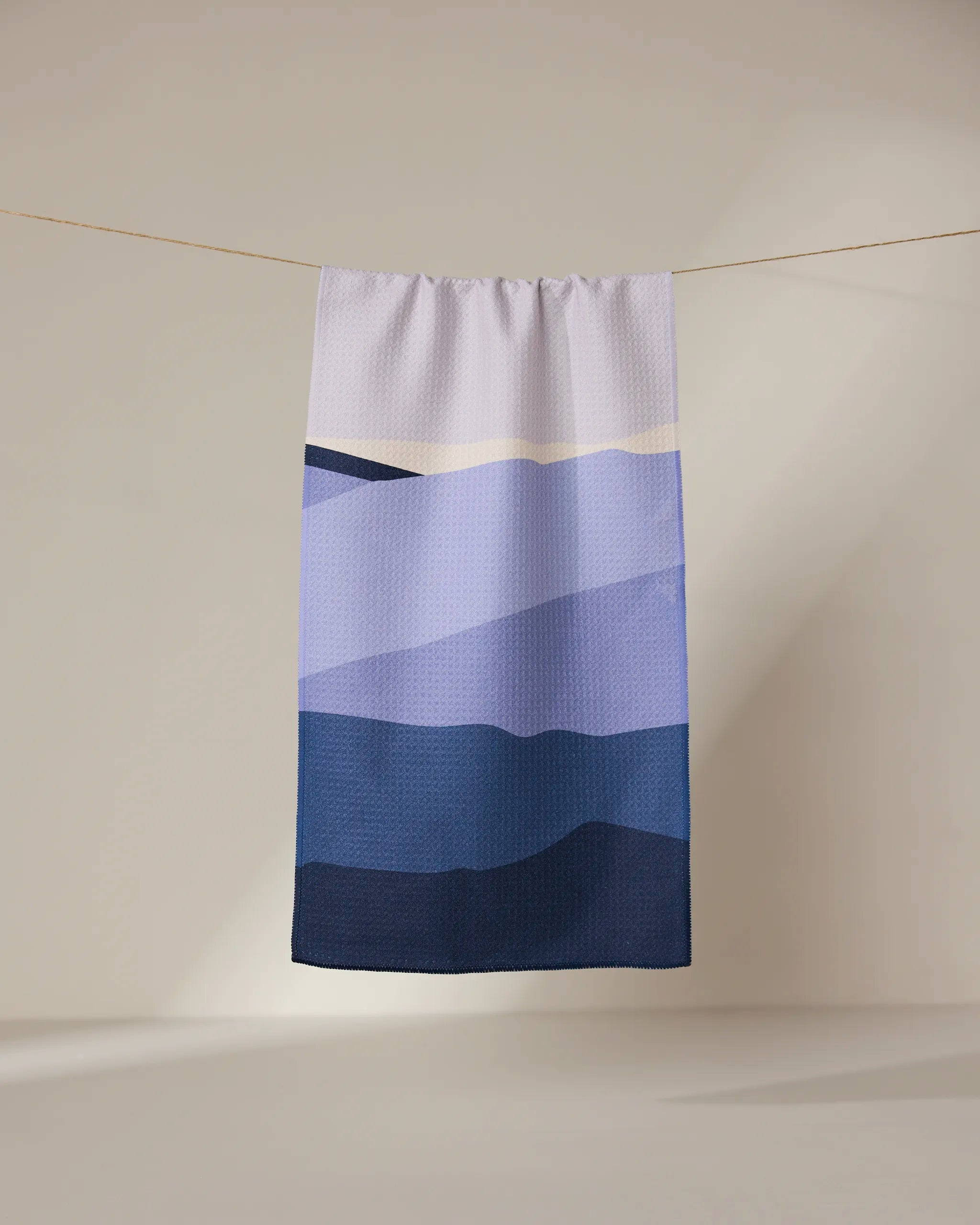 The GEOMETRY Mountainous rectangular towel, ultra-absorbent and adorned with blue and purple mountain-inspired gradients, hangs from a string against a neutral background.