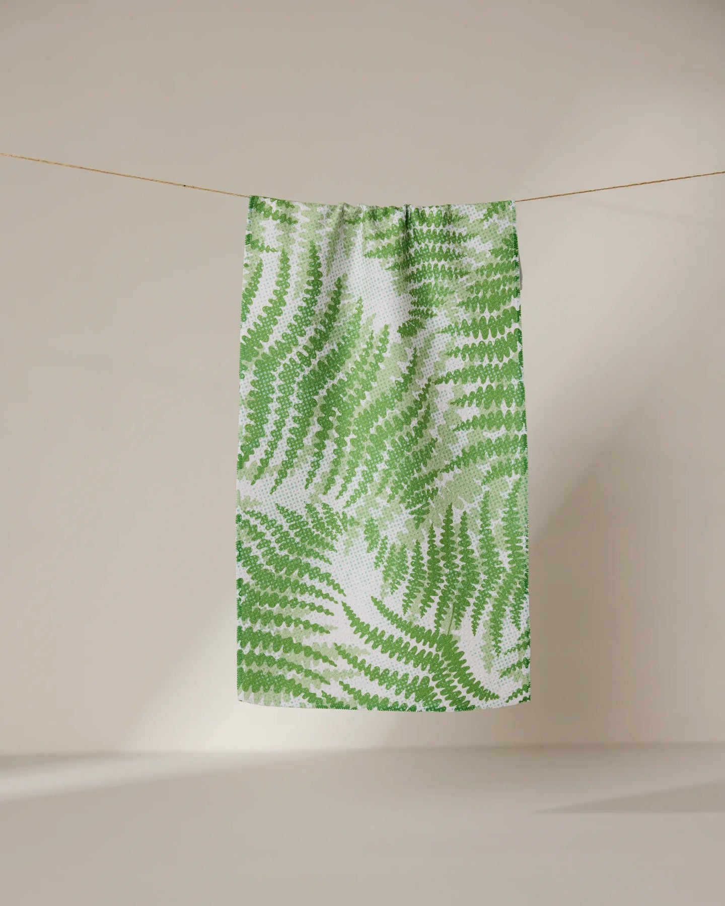 The GEOMETRY Swaying Leaf towel, featuring a green fern pattern, dries quickly and hangs on a clothesline against a neutral backdrop.