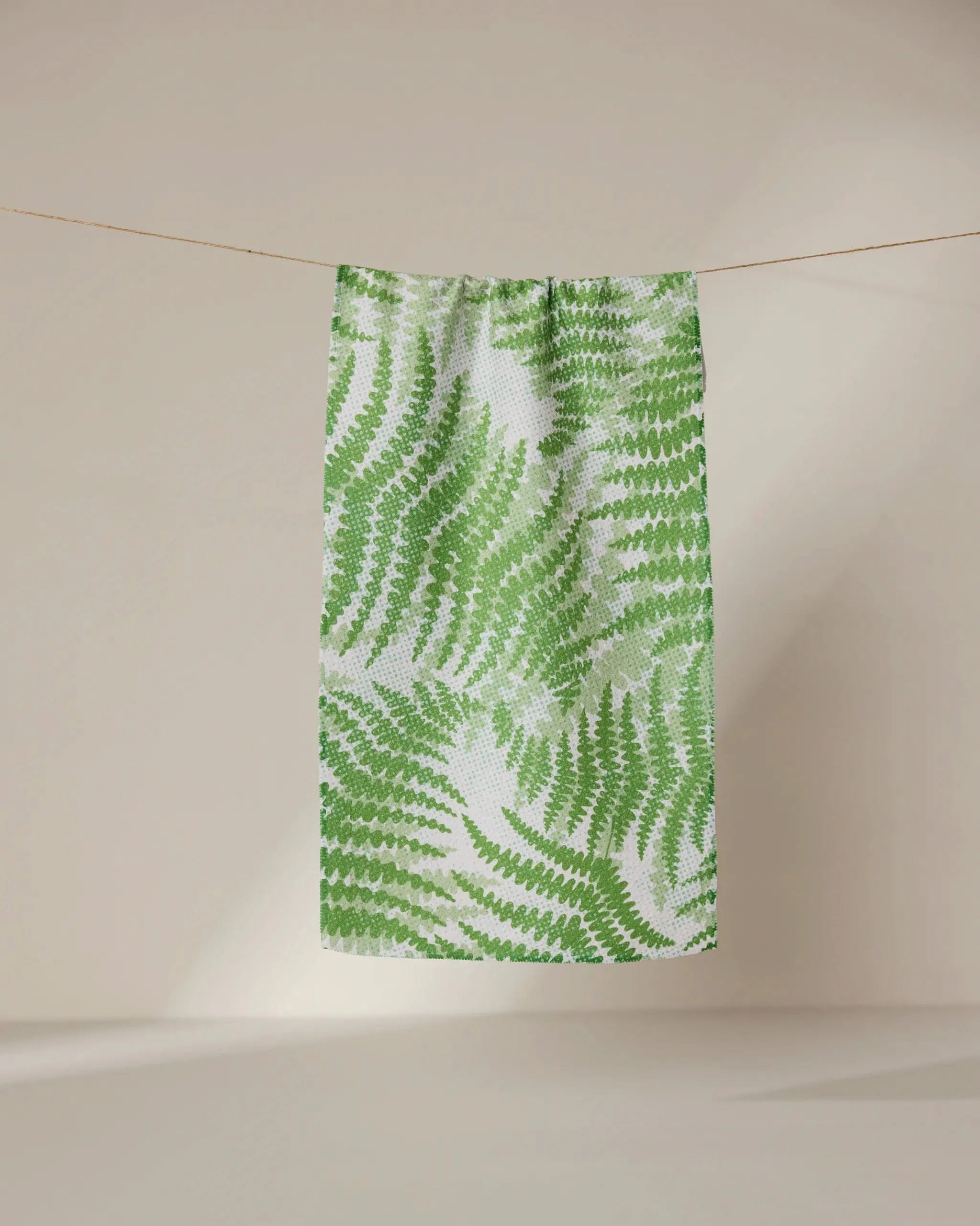 The GEOMETRY Swaying Leaf towel, featuring a green fern pattern, dries quickly and hangs on a clothesline against a neutral backdrop.