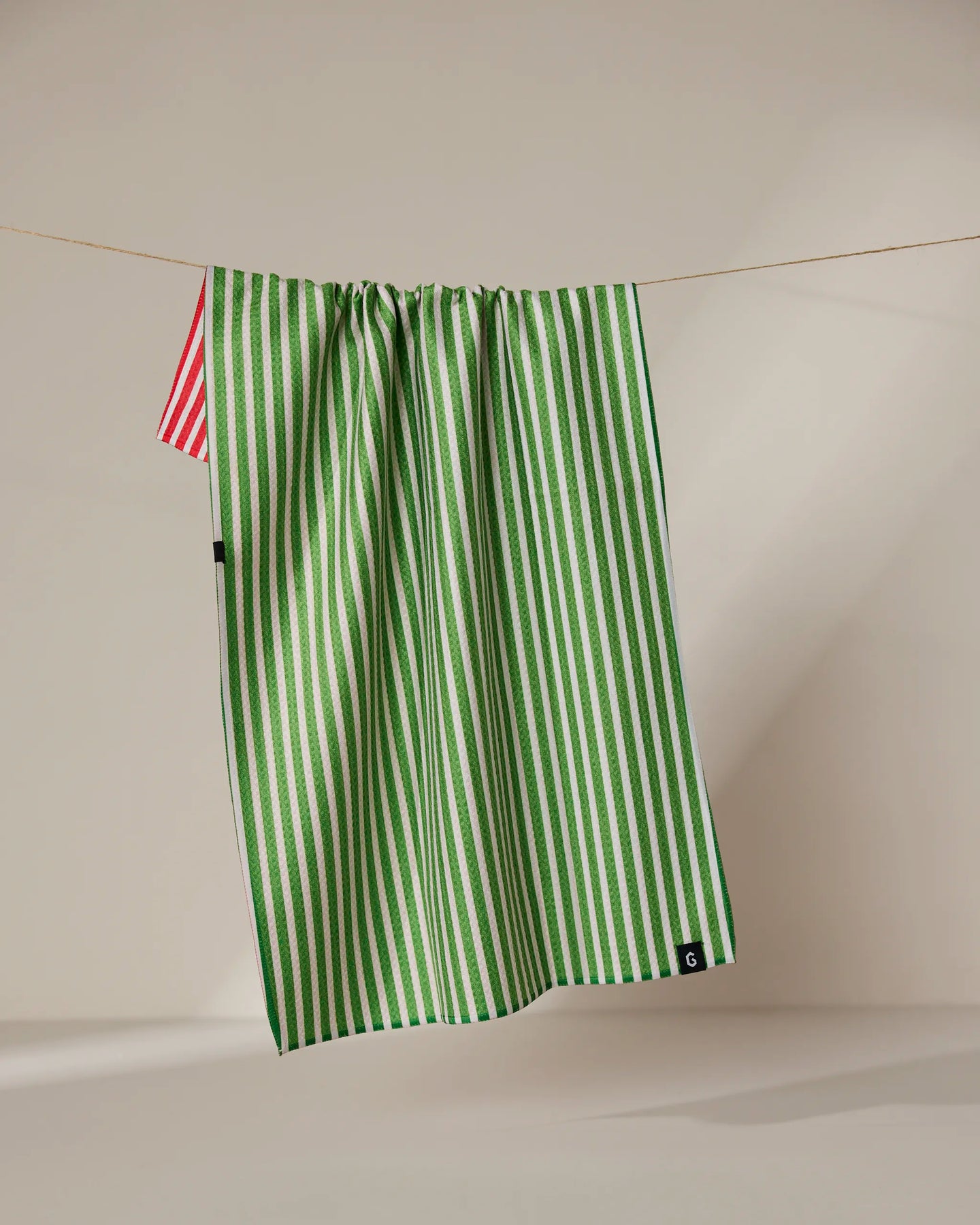 The Geometry Red And Green double-sided tea towel hangs from a clothesline, featuring green and white stripes with a hint of red and white stripes visible against a neutral background.