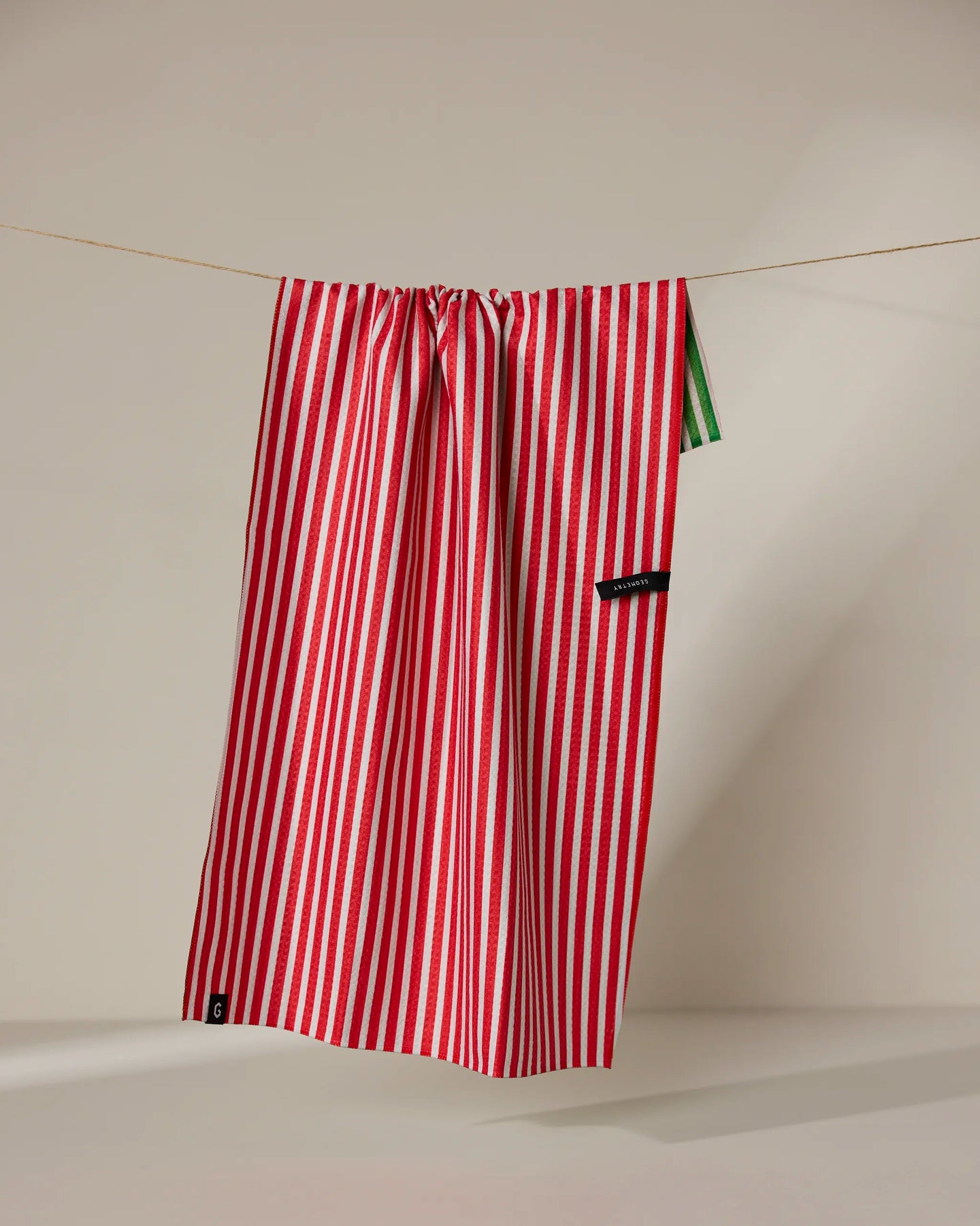 The Geometry Red And Green tea towel, featuring red and white vertical stripes and quick-drying fabric, hangs from a string against a neutral background. A small black label and a touch of green are visible on one side.