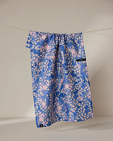 The Geometry Lav tea towel, featuring blue and pink abstract shapes and a small black tag, hangs on a clothesline against a plain light background. Its quick-drying fabric displays the brands signature modern pattern.