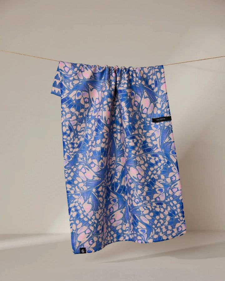 The Geometry Lav tea towel, featuring blue and pink abstract shapes and a small black tag, hangs on a clothesline against a plain light background. Its quick-drying fabric displays the brands signature modern pattern.