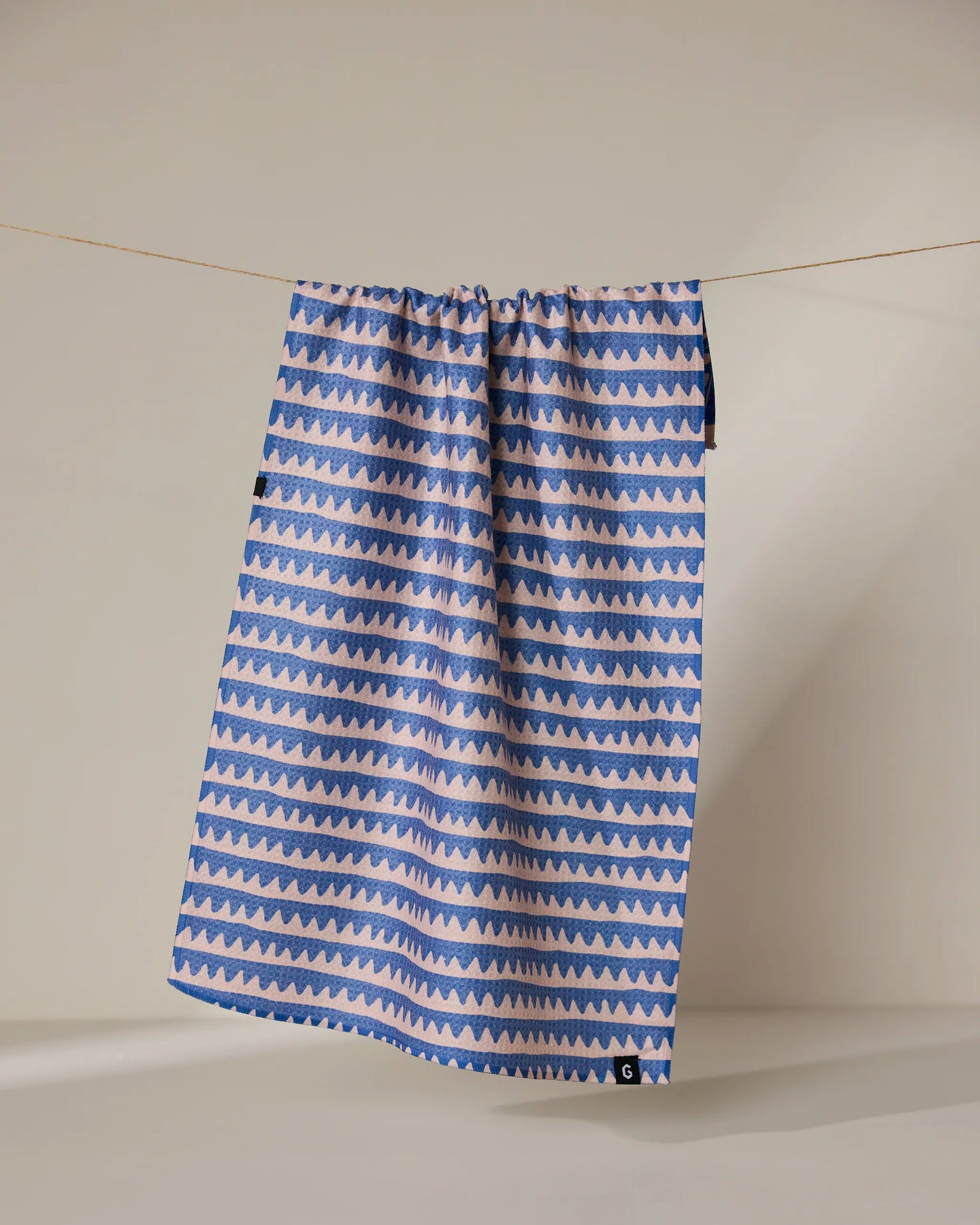 The Geometry Lav tea towel features double-sided alternating blue and light pink zigzag stripes, shown hanging on a clothesline against a neutral background.