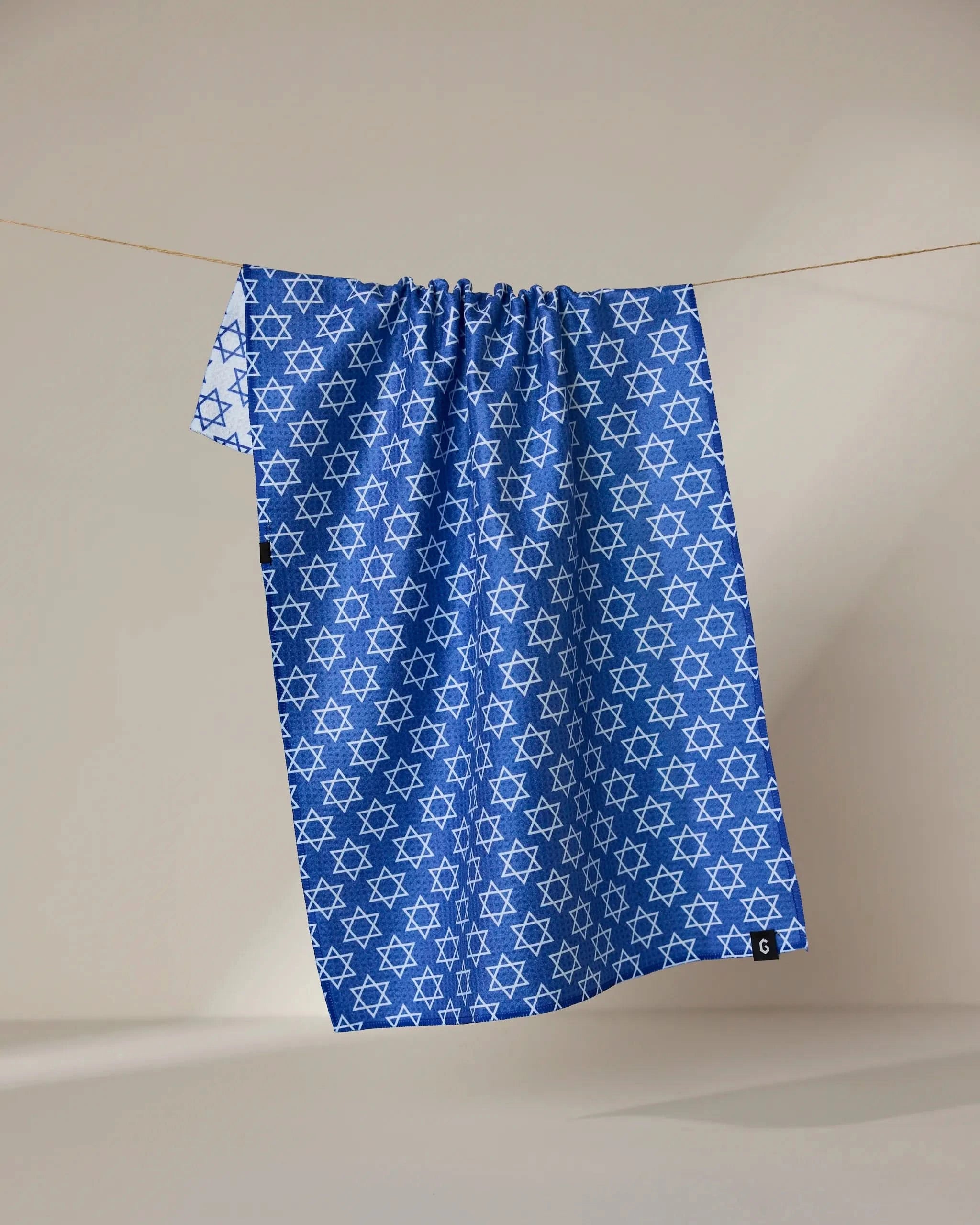 A blue recycled Star Of David kitchen towel by Geometry, featuring a repeating white Star of David pattern, hangs on a clothesline against a neutral background.
