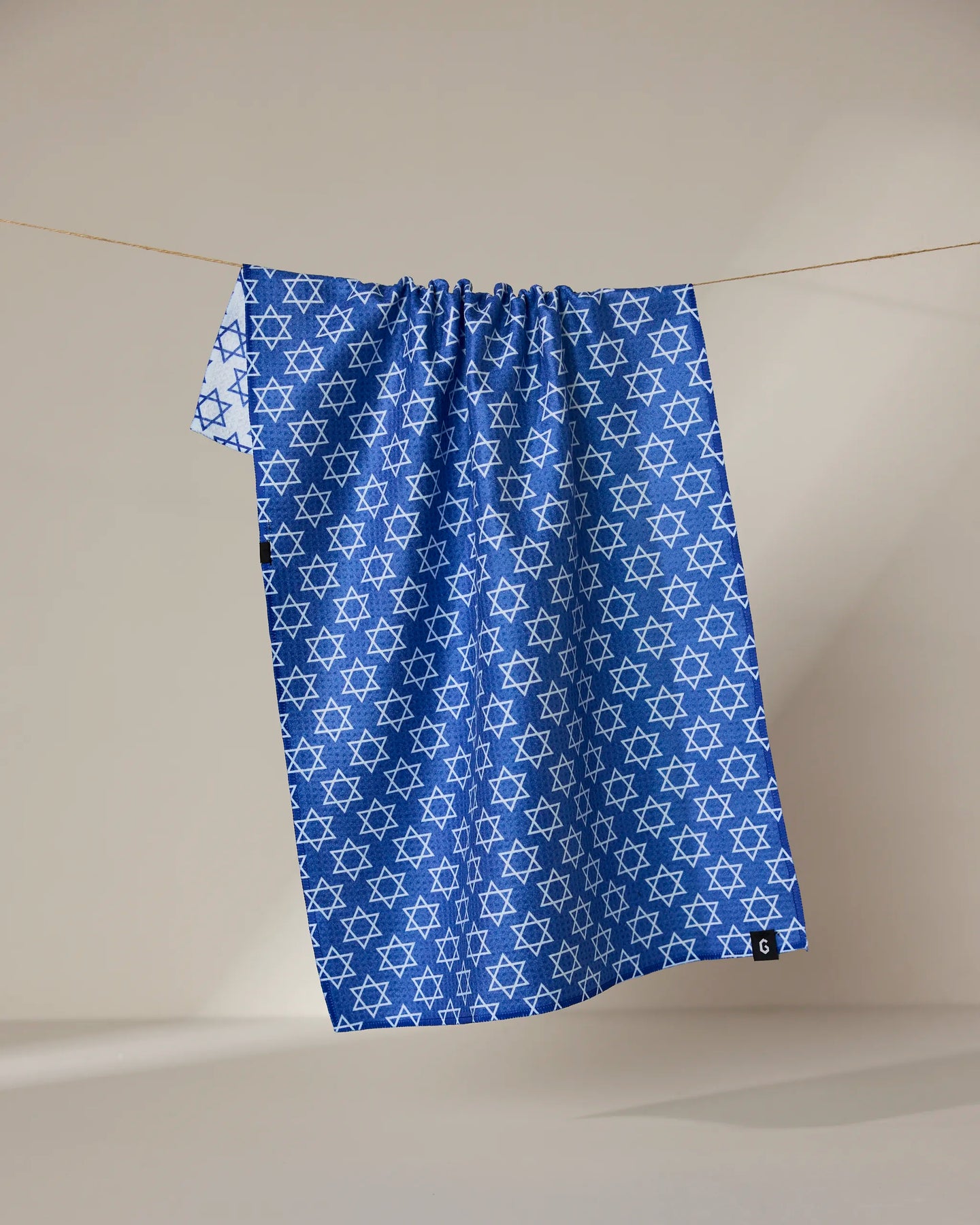 A blue recycled Star Of David kitchen towel by Geometry, featuring a repeating white Star of David pattern, hangs on a clothesline against a neutral background.