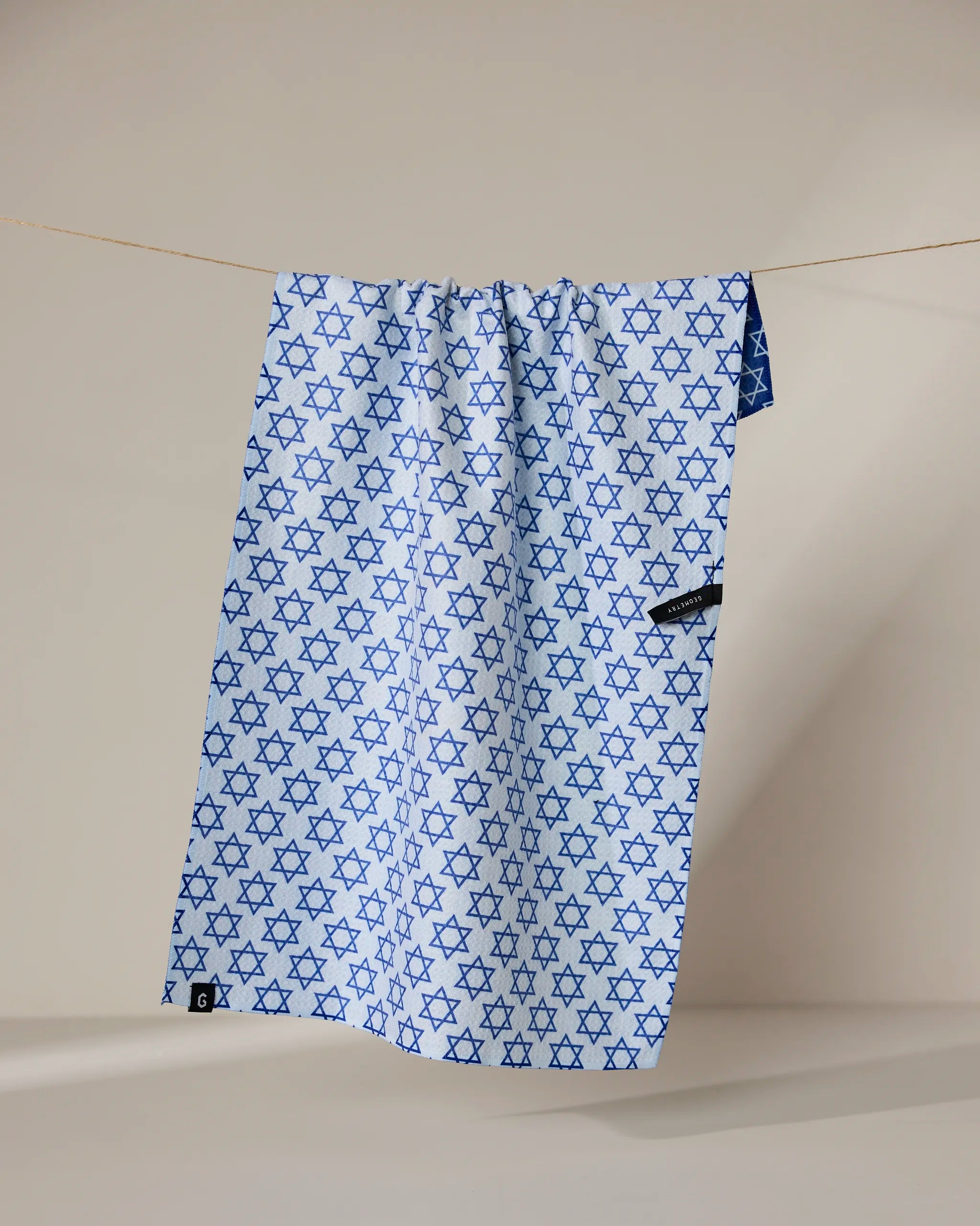 The Geometry Star Of David kitchen towel features a blue Star of David pattern on light blue, hangs on a clothesline with a visible black tag, and is made from recycled materials for quick drying.