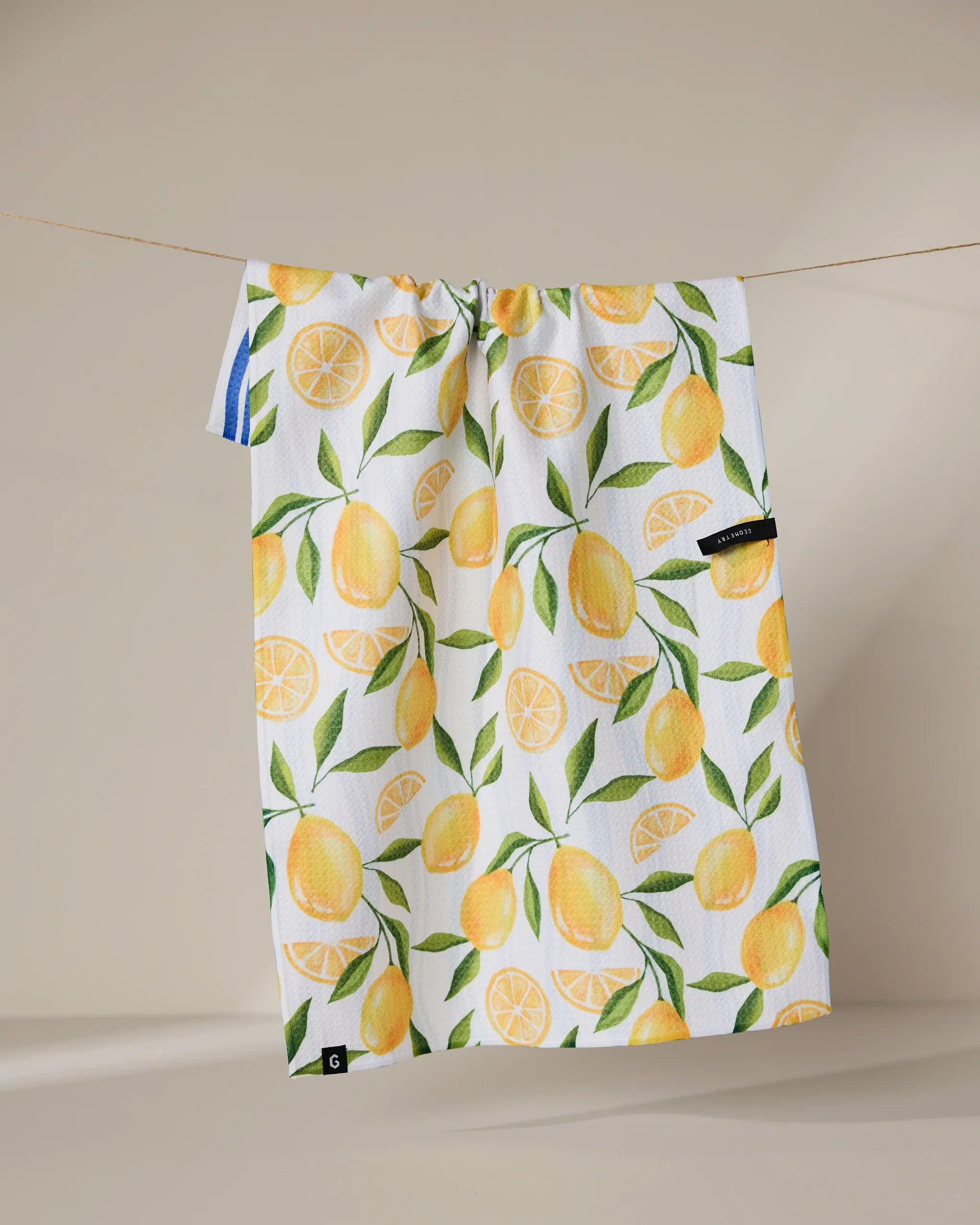 The Geometry Lemon Bliss towel, white and quick-drying with a vibrant yellow lemon and green leaf pattern, hangs on a clothesline against a neutral background.