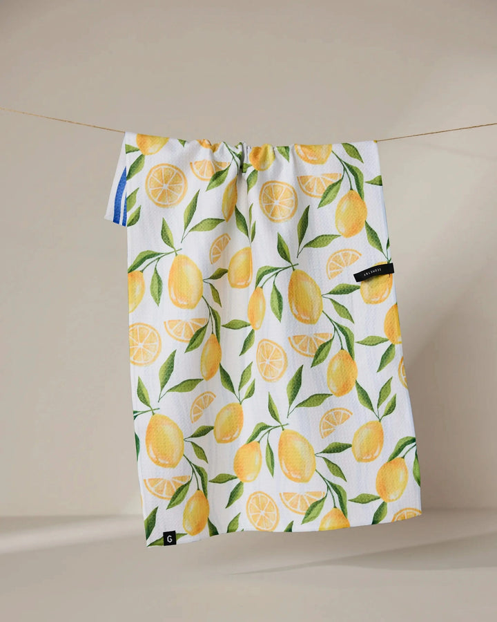 The Geometry Lemon Bliss towel, white and quick-drying with a vibrant yellow lemon and green leaf pattern, hangs on a clothesline against a neutral background.