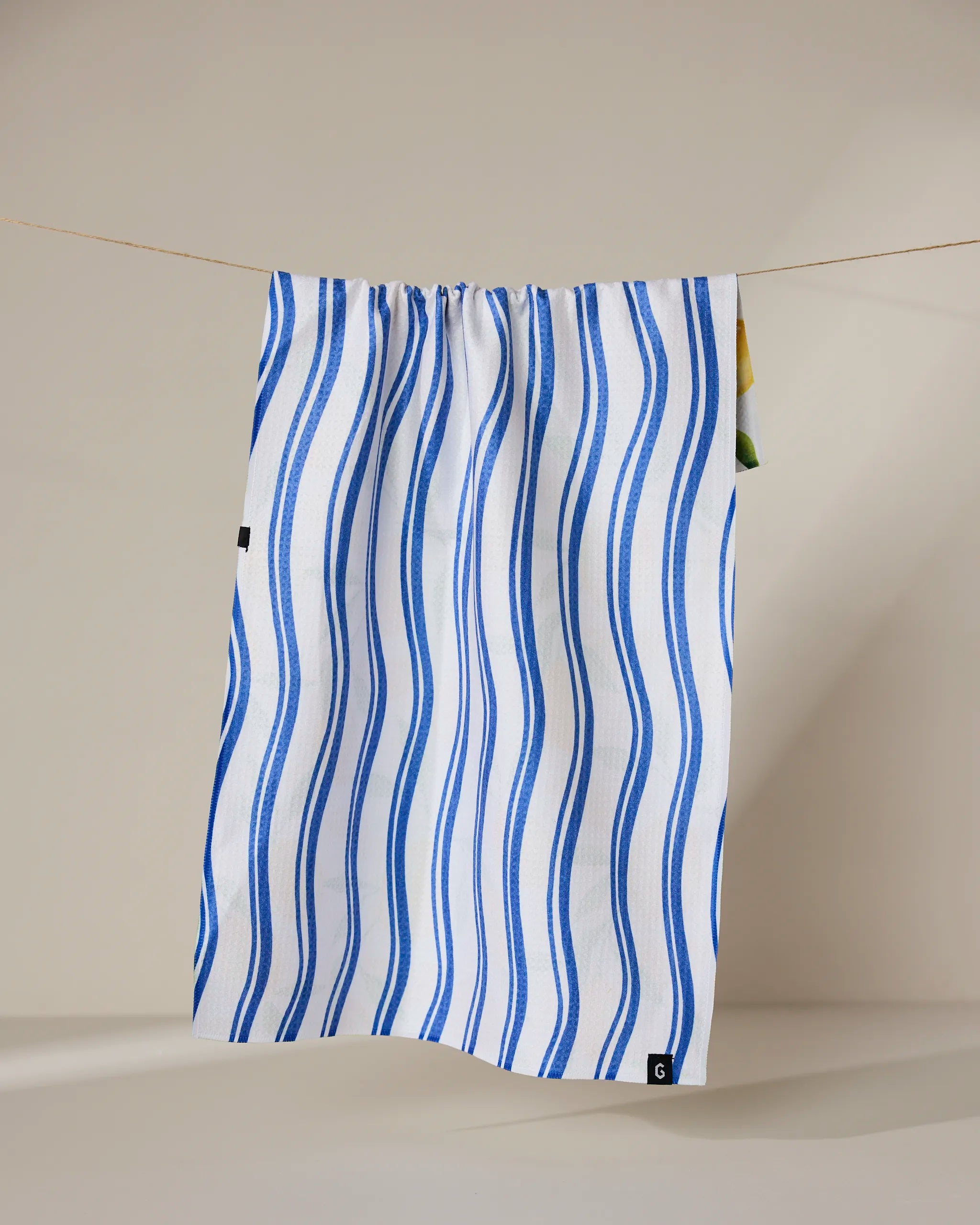 The Geometry Lemon Bliss tea towel, featuring wavy blue stripes on white fabric, hangs from a clothesline against a simple light background.
