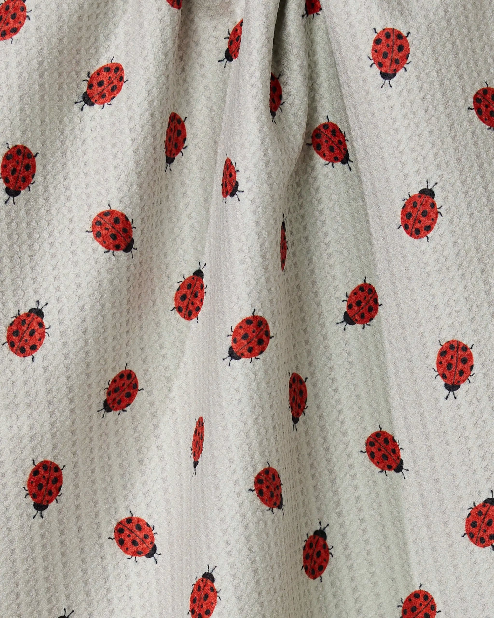 A close-up of the Geometry Lady tea towel, made from quick-drying, light-colored textured fabric with a repeating red and black ladybug pattern. The material is gently gathered, creating soft folds and shadows.