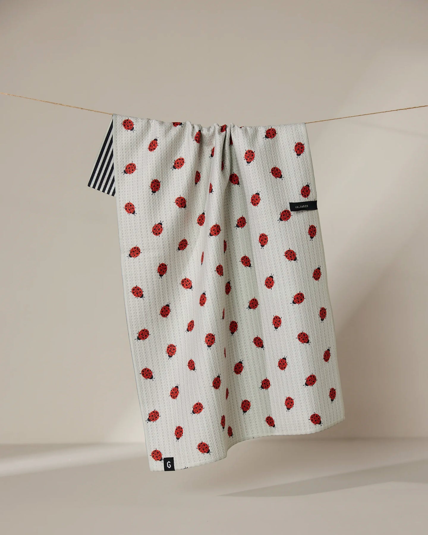 The Geometry Lady kitchen towel, made from recycled materials, features a small red strawberry pattern, black and white striped edge, black label, and hangs on a thin clothesline against a softly lit neutral background.