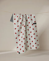 The Geometry Lady kitchen towel, made from recycled materials, features a small red strawberry pattern, black and white striped edge, black label, and hangs on a thin clothesline against a softly lit neutral background.