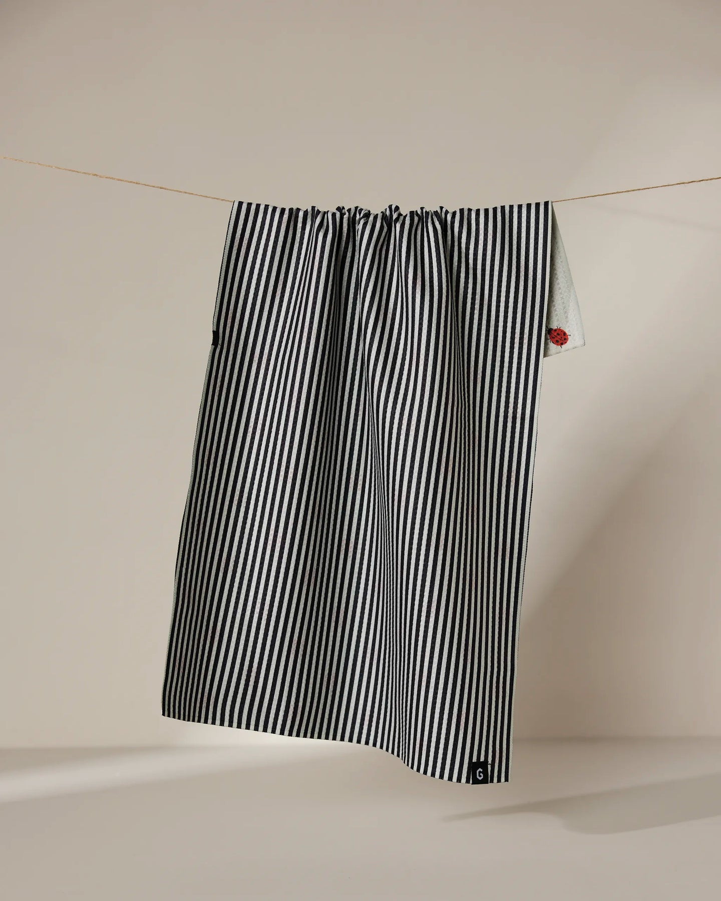 The Geometry Lady tea towel features black and white vertical stripes and a small red patch near the top right corner. It hangs double-sided on a thin clothesline against a neutral background.