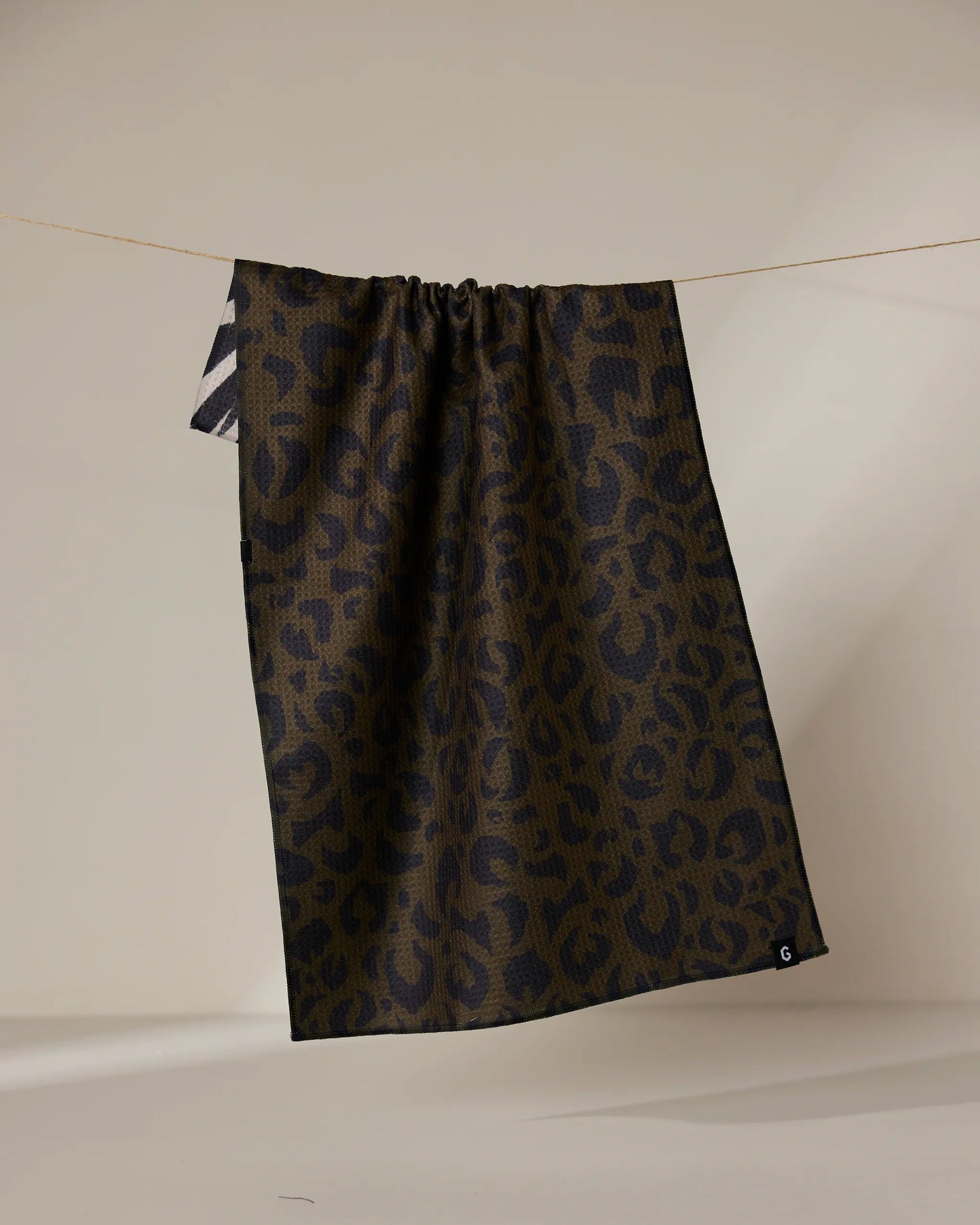 A dark brown and black leopard print Geometry Safari Absorbent Quick-Dry Tea Towel hangs on a thin clothesline against a neutral background, featuring a small white G in one corner and double-sided design.